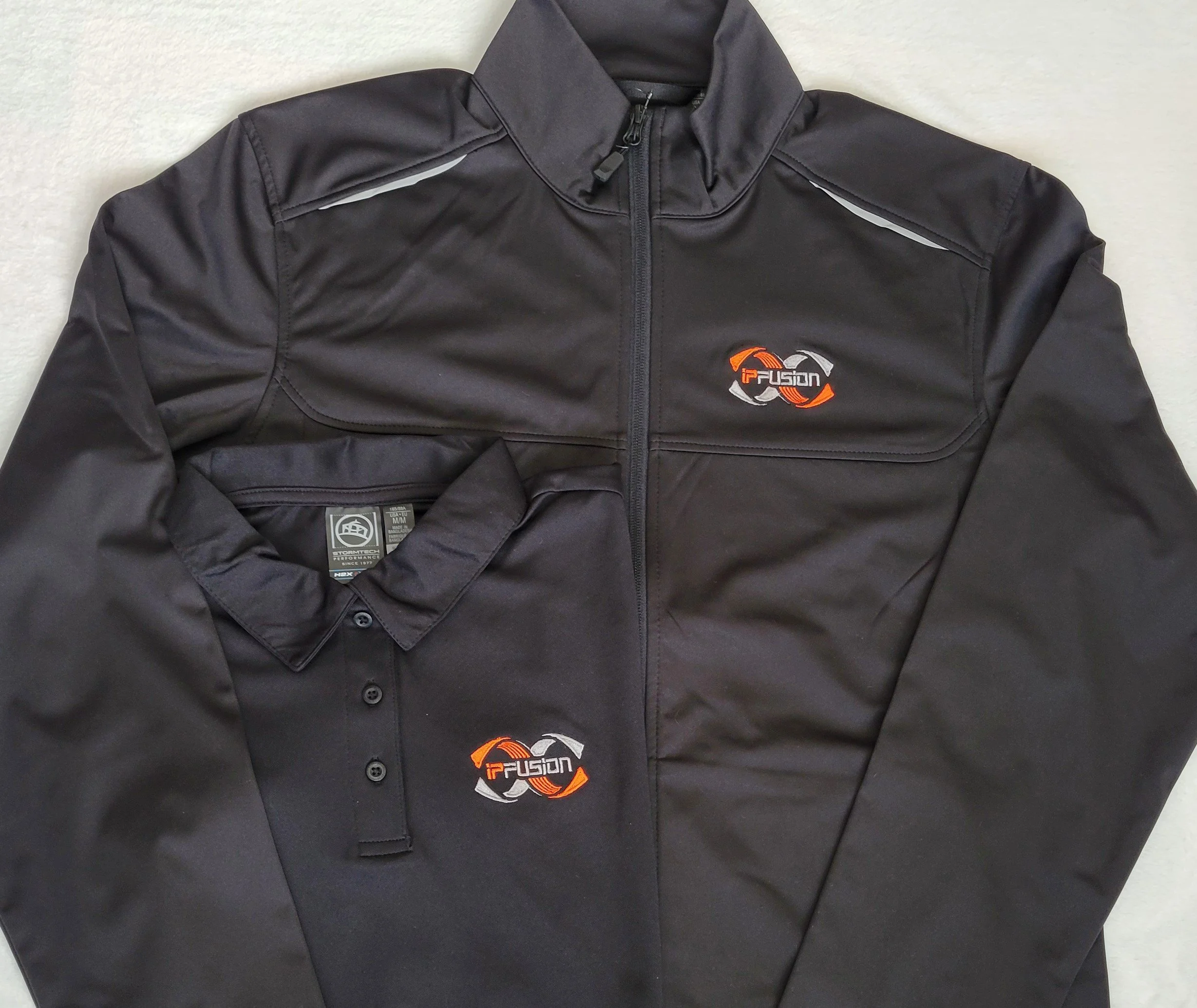 A black athletic jacket with a full front zipper and the 'ipFusion' logo on the chest, placed over a black polo shirt also with the 'ipFusion' logo.