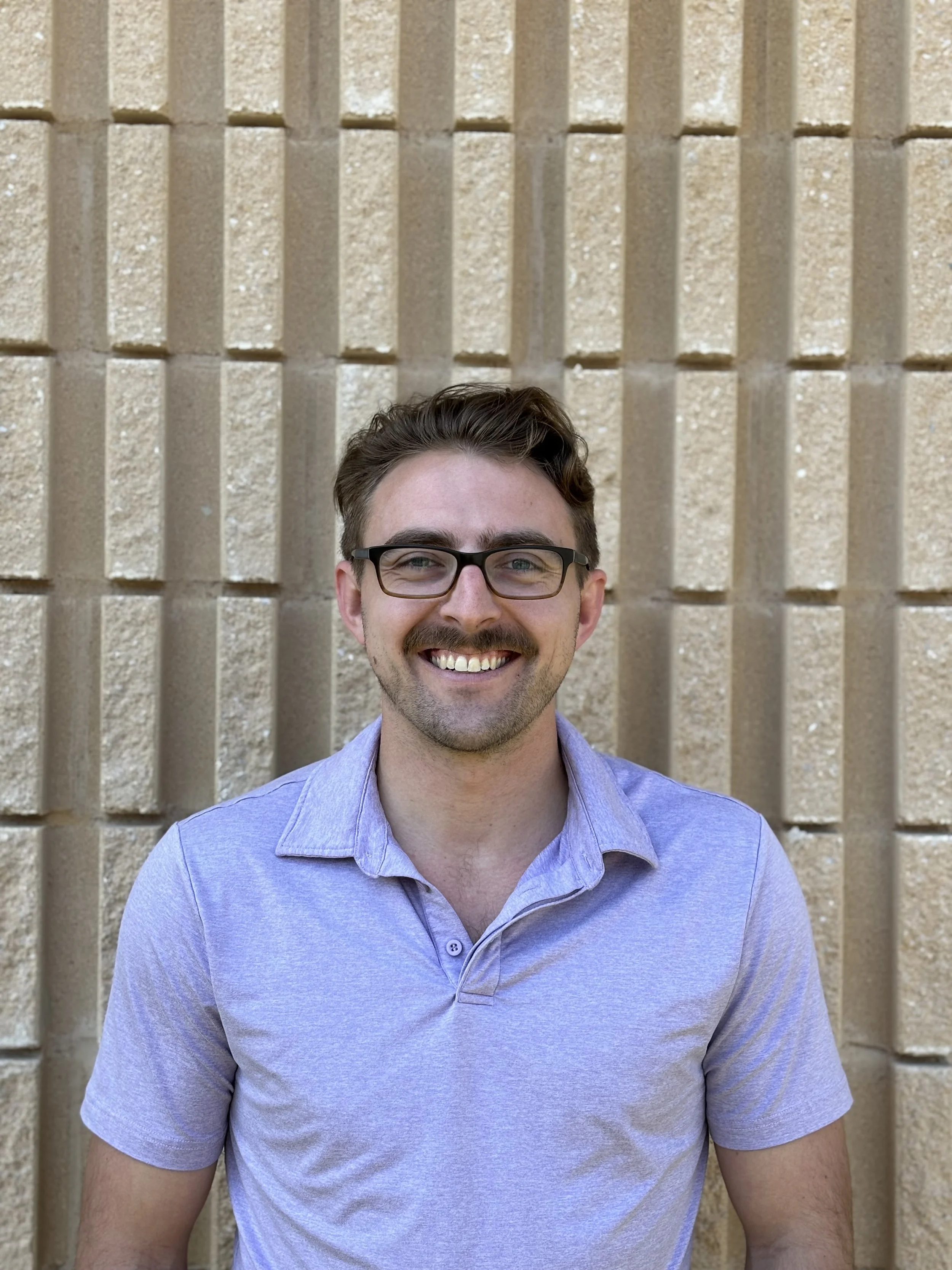 Smiling person with glasses in front of a textured beige brick wall, wearing a light purple collared shirt.