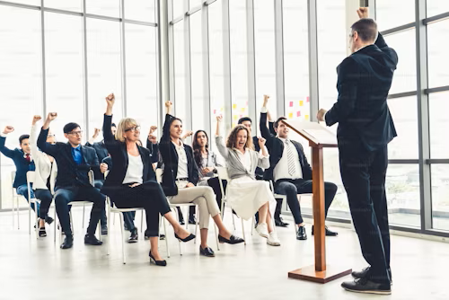 A man giving a presentation to a group of diverse people in a modern office with large windows. The audience is raising their hands.