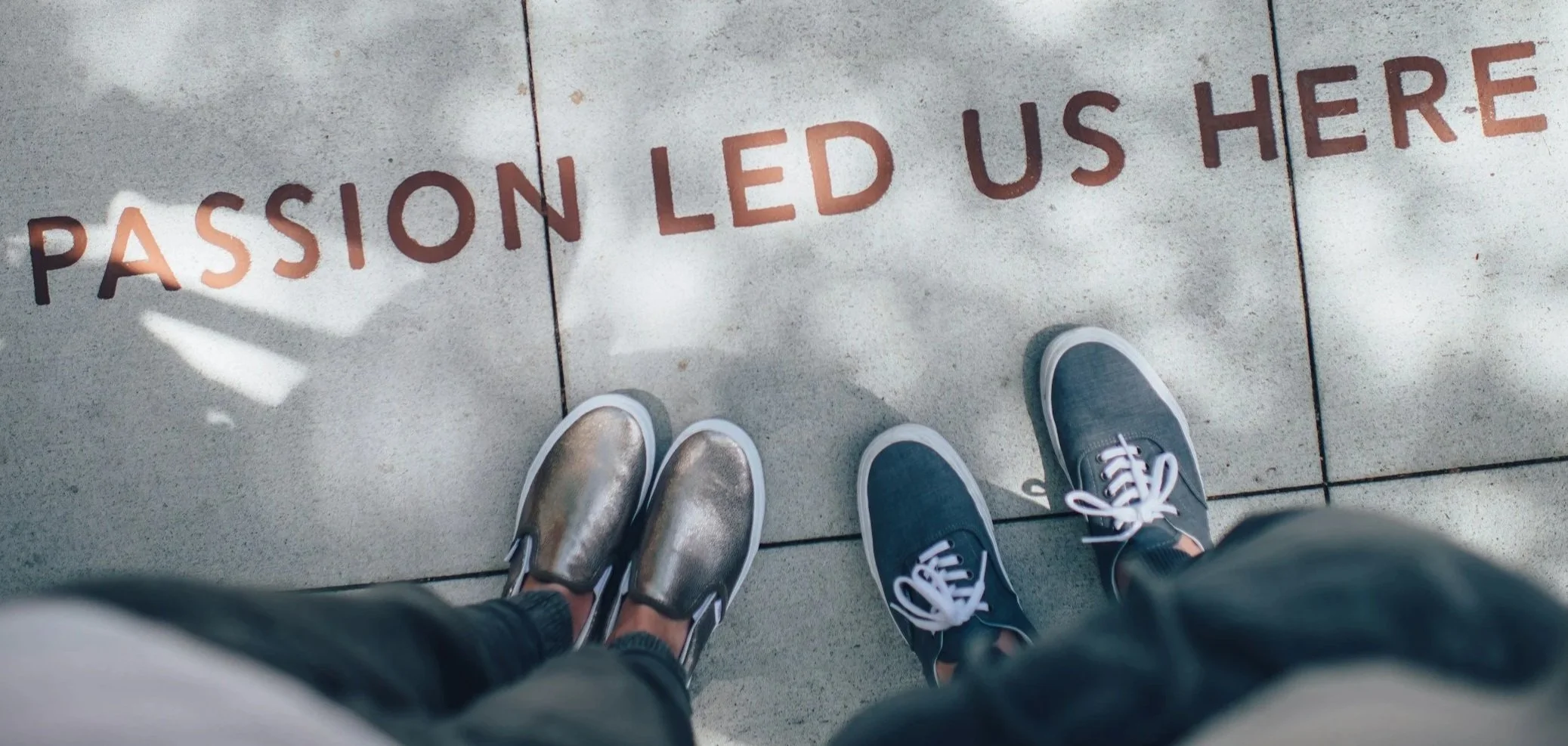 View of two people's feet on a sidewalk, with the words 'Passion Led Us Here' written in red on the ground.
