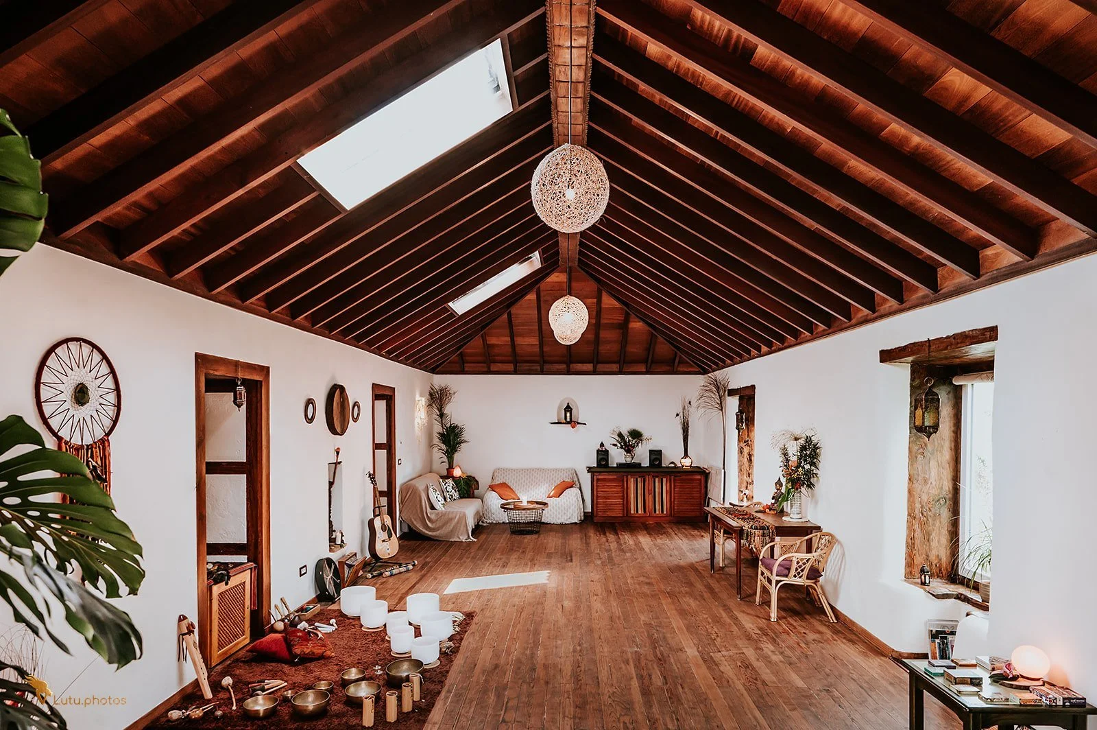 A spacious living room with a high vaulted wooden ceiling, skylights, and hardwood flooring. It is decorated with various plants, guitars, candles, and vintage furniture, creating a cozy and bohemian atmosphere.