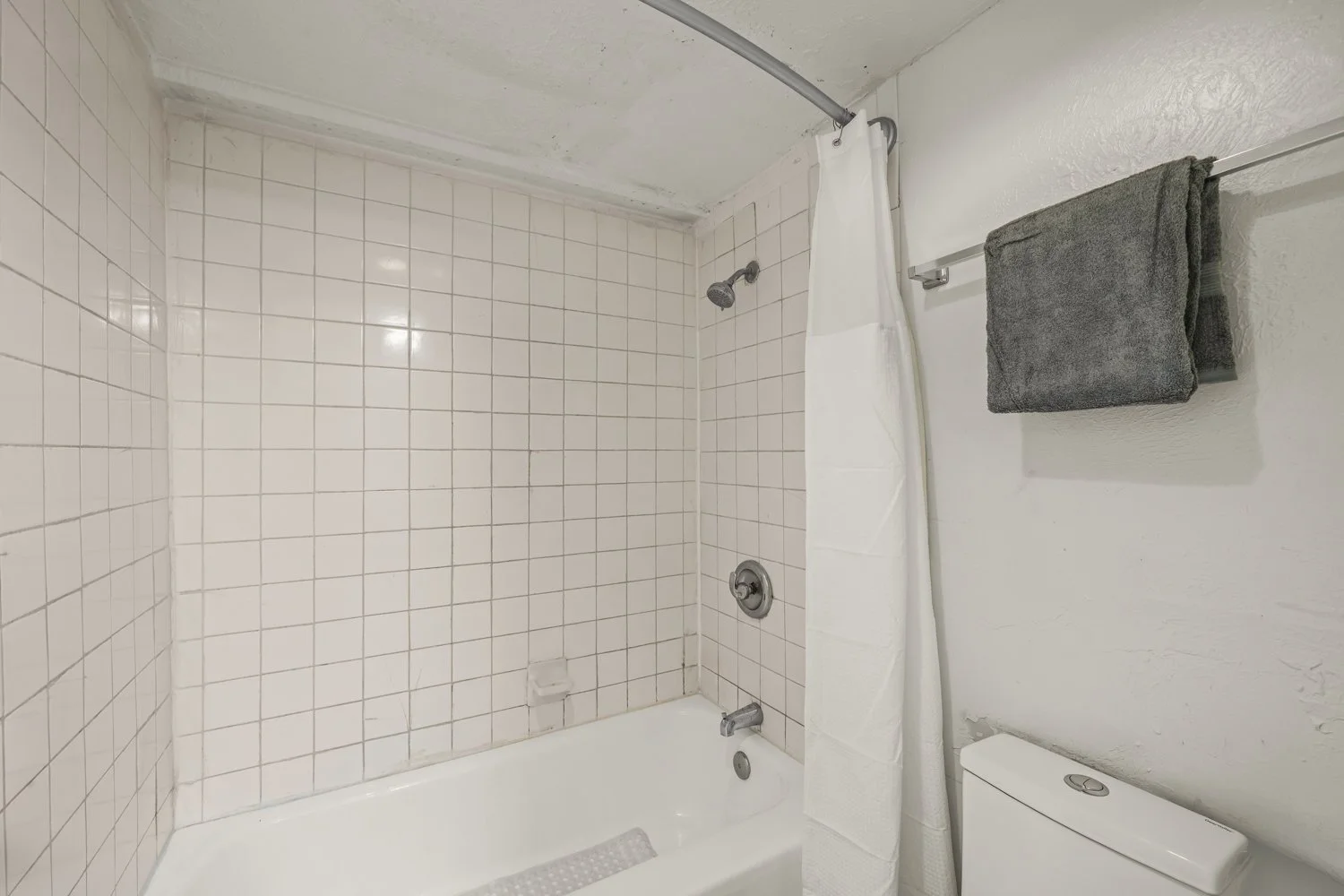 Bathroom with a tiled shower area, shower head, white tub, towel rod with a gray towel, and part of a toilet.