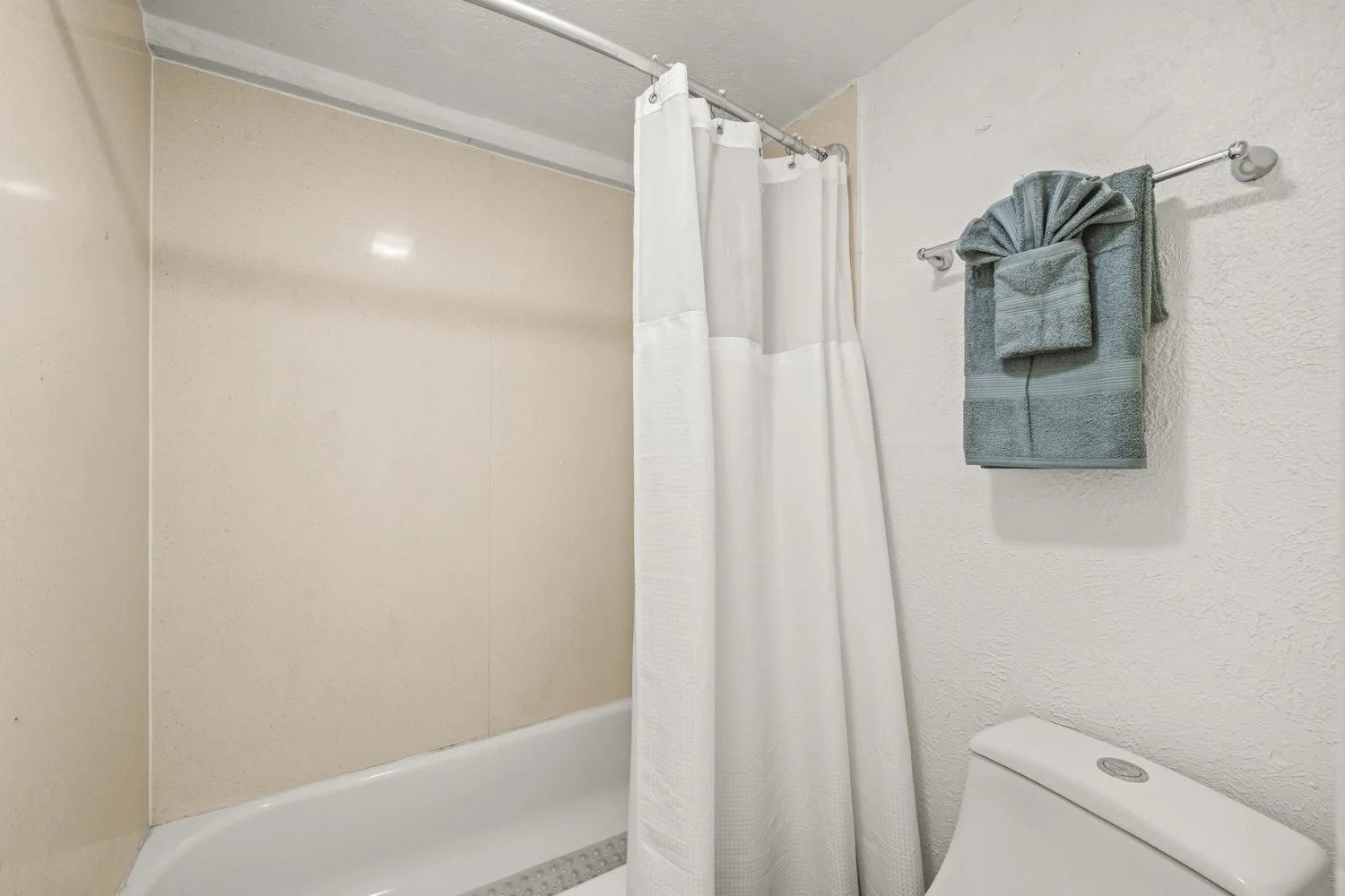 Bathroom with a shower and a toilet, beige and white walls, a white shower curtain, and gray towels hanging on a wall-mounted towel rod.