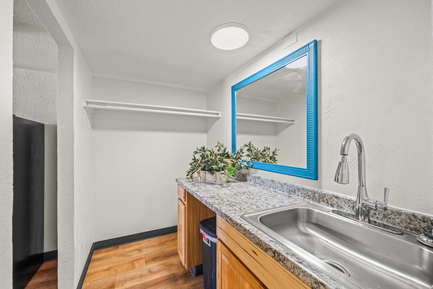 A small kitchenette with a granite countertop, wooden cabinets, a stainless steel sink, a blue framed mirror, a potted plant, and a white shelf on the wall.