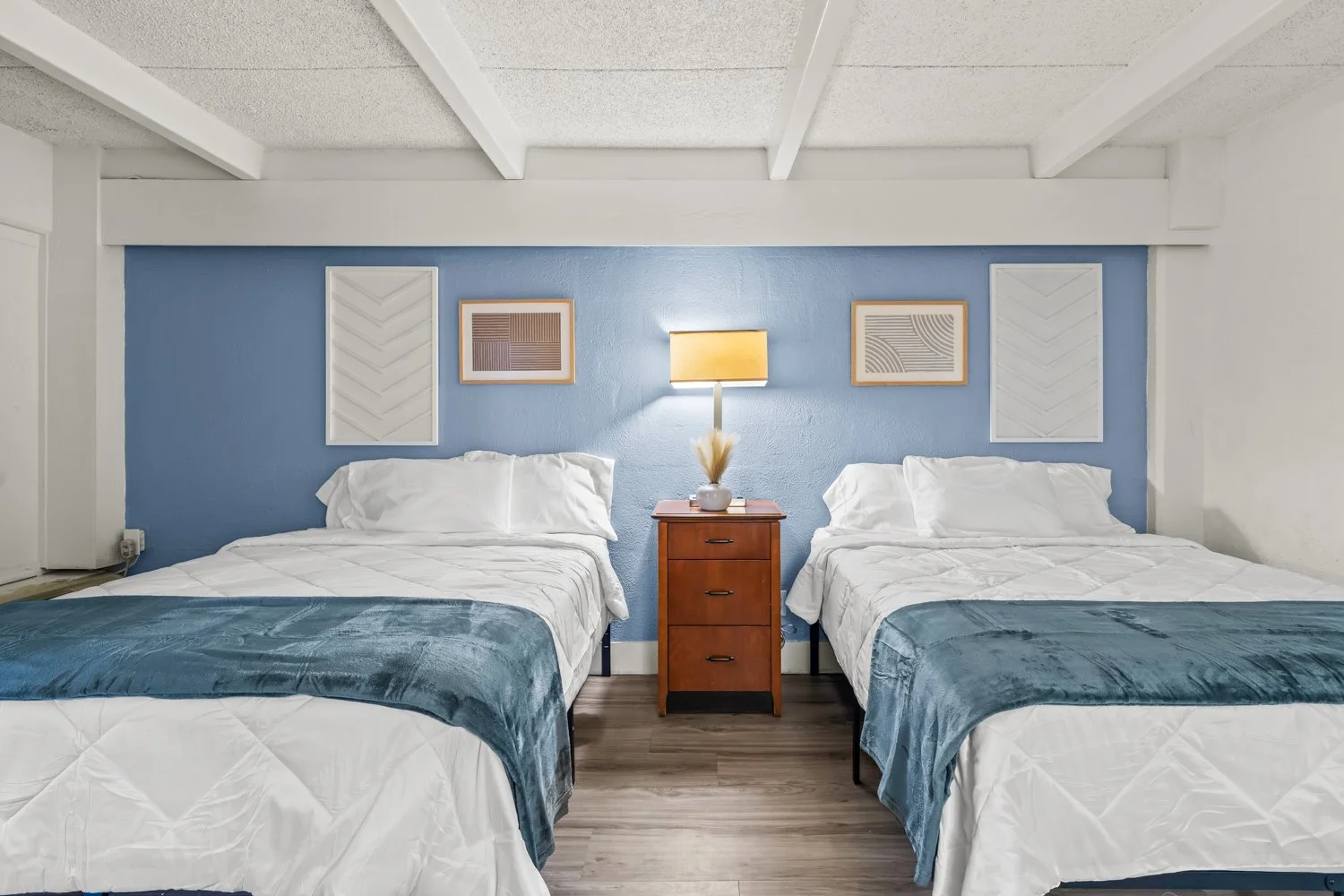 A bedroom with two double beds separated by a wooden nightstand, blue accent wall, framed artwork, and a table lamp.