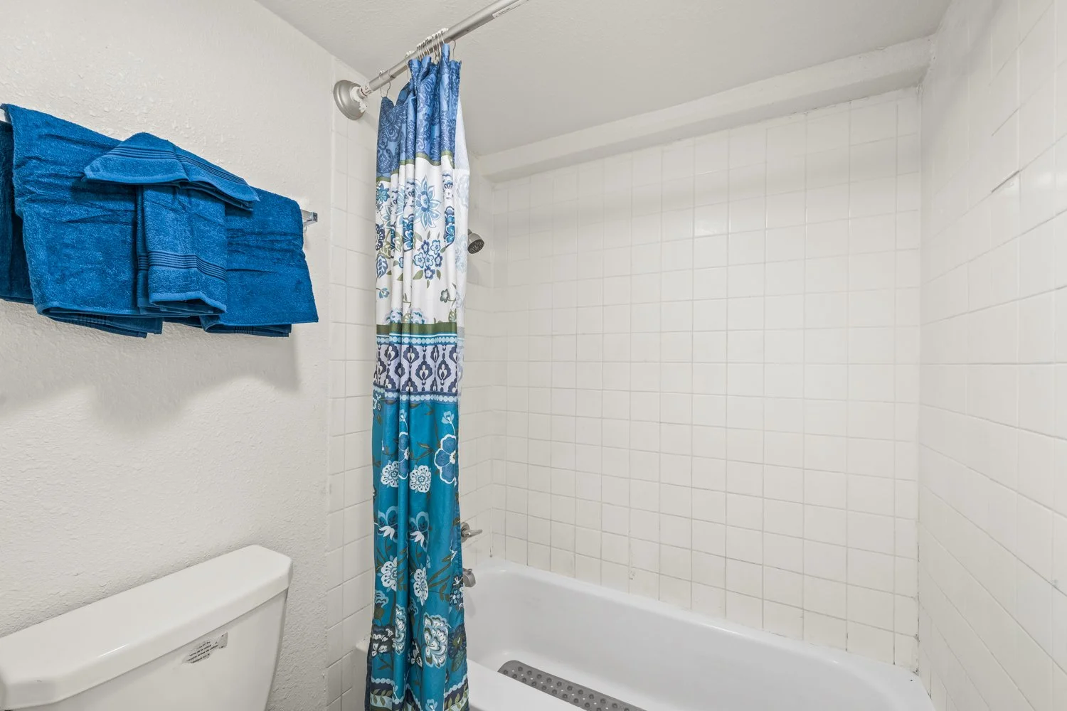 Bathroom shower area with a blue floral shower curtain, white tiled wall, and three blue towels on a wall-mounted towel rack.