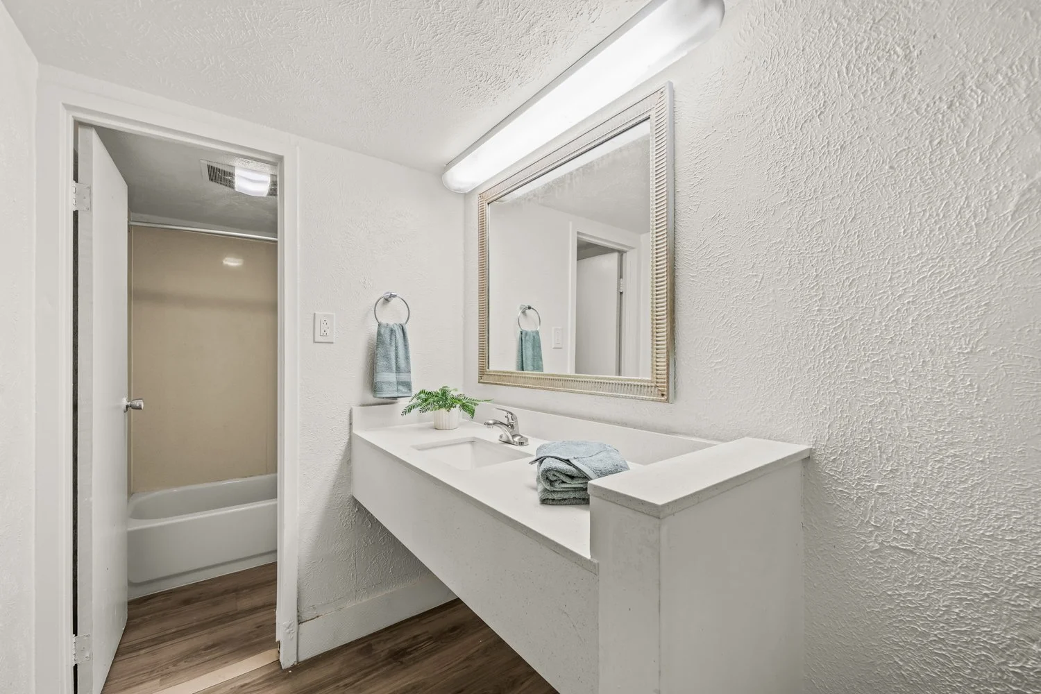 A small bathroom with a white vanity, large mirror, and a bathtub/shower combination. The room has white textured walls, wood-look flooring, and a towel holder with a blue towel.
