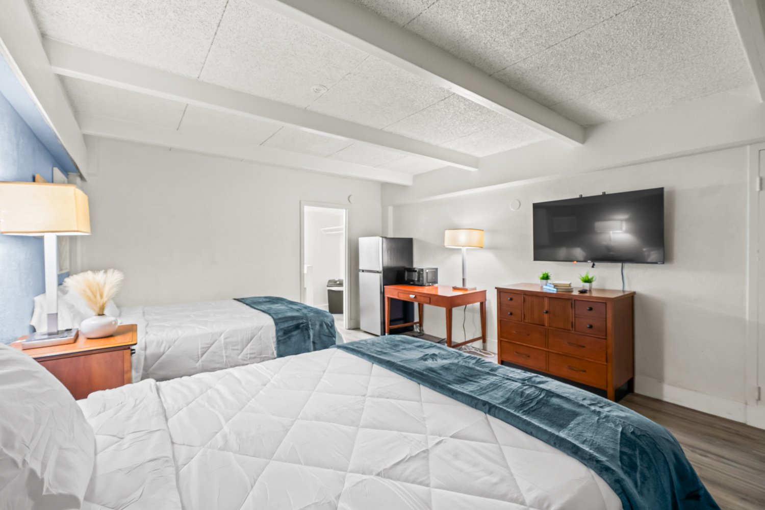 Two beds in a room with white quilted bedding and blue throws, a wooden nightstand with a lamp and decorative vase, a wooden dresser with small plants and books, a mounted flat-screen TV, a red desk with a microwave and coffee maker, and a refrigerat