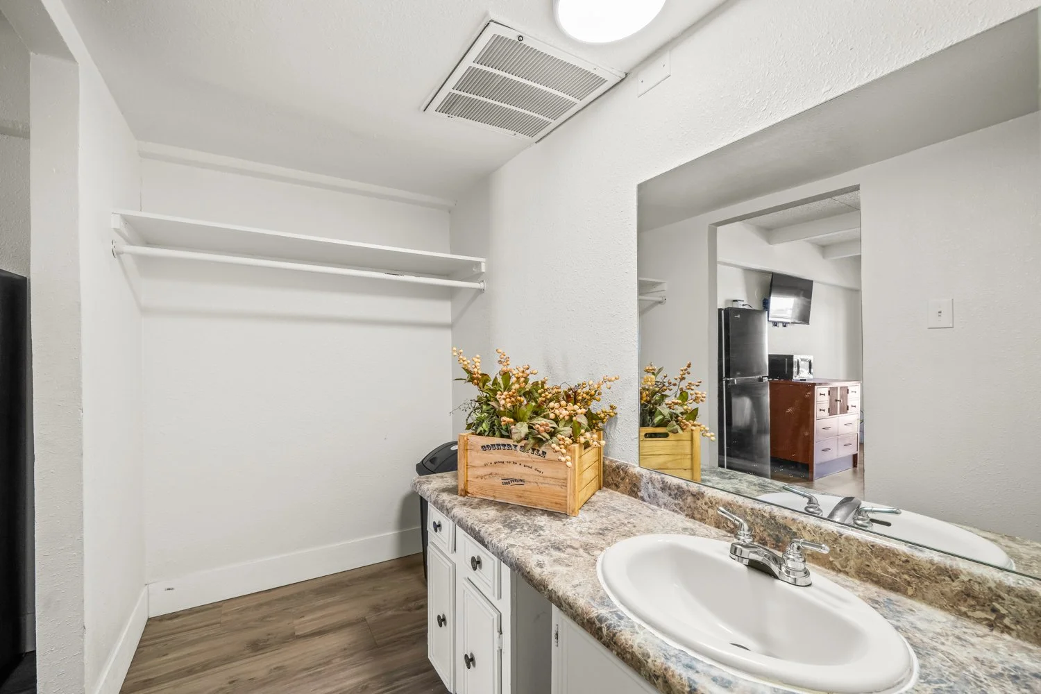 Empty bathroom vanity with a sink, a large mirror, a potted plant in a wooden box, and an open shelving area above. The mirror reflects part of the kitchen with a refrigerator and a wooden cabinet.