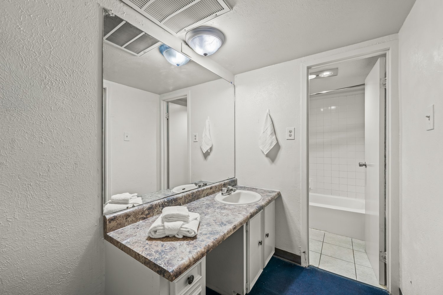 Bathroom with white walls, a granite countertop with a sink, a large mirror, and a towel hanging on the wall. There is a door leading to a bathtub/shower area with white tile walls.
