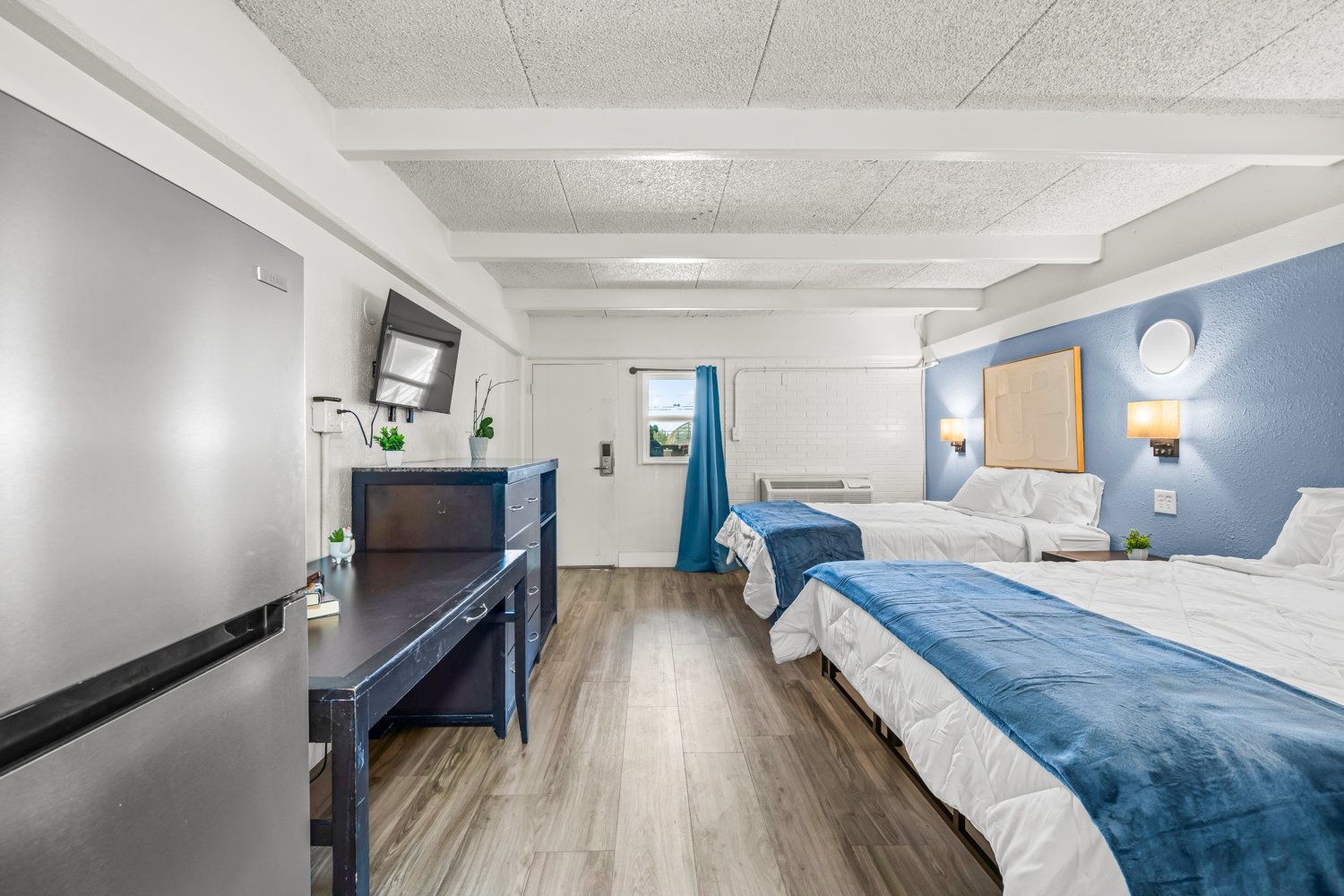 A hotel room with two beds, one with a blue blanket, a small nightstand, and wall-mounted lamps, with a blue accent wall and a white brick section near the window, a flat-screen TV on a blue dresser, and a kitchenette with a refrigerator and countert