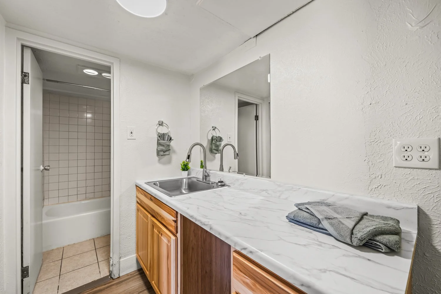 Kitchen with white marble countertop, wooden cabinets, a stainless steel sink, and a large mirror on the wall. Towels hang on ring holders, and small potted plants sit on the counter. An open door leads to a shower with beige tiles.