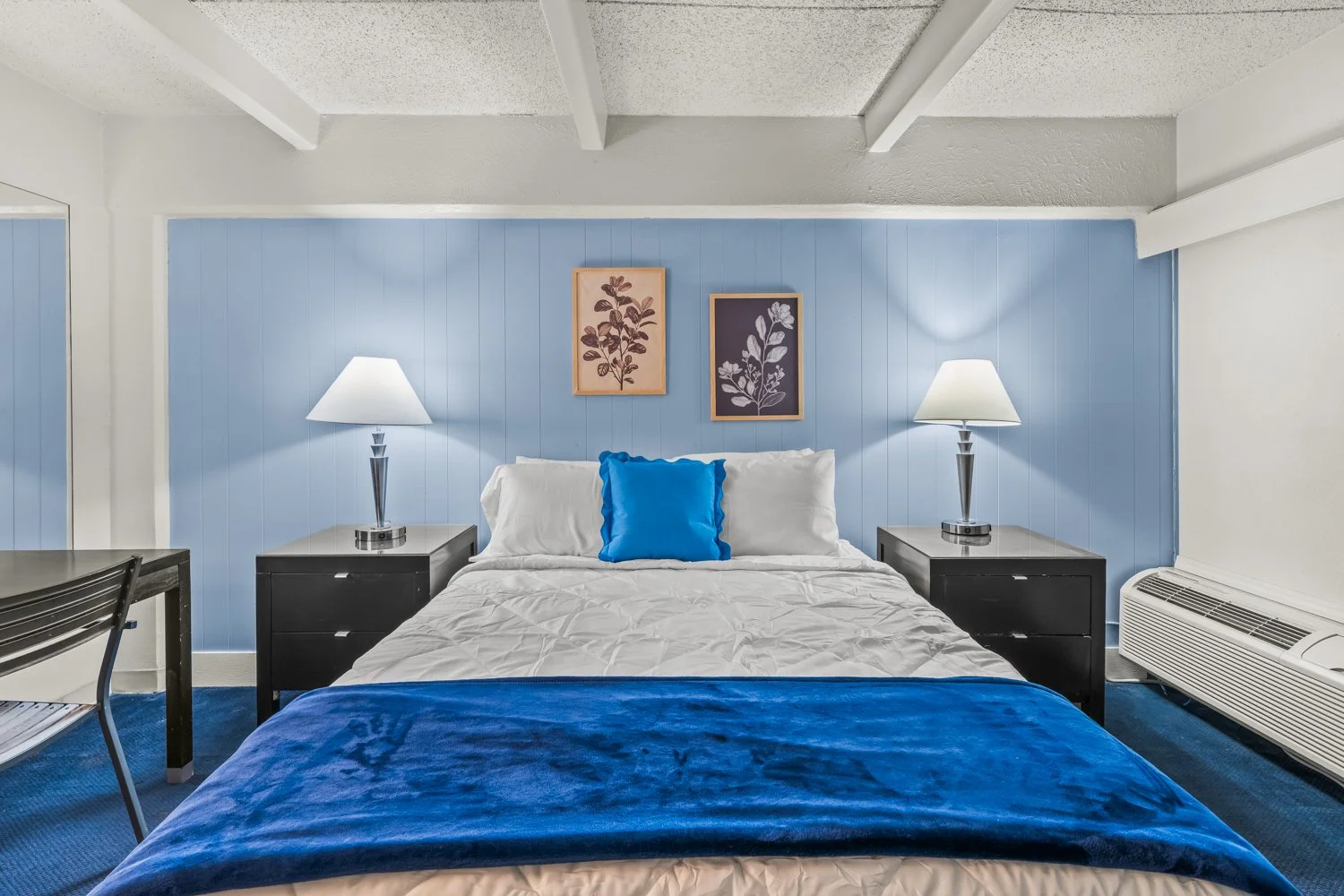 Hotel room with a bed, two nightstands with lamps, artwork above the bed, dark furniture, blue accents, and a window air conditioner.