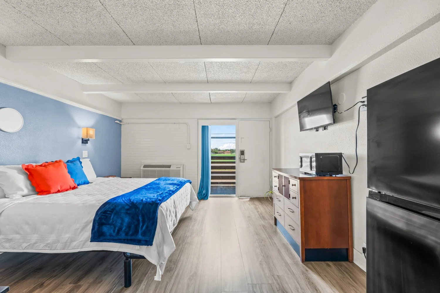 Hotel room with a bed featuring red and blue pillows, a blue throw blanket, a wooden dresser with a microwave and a small TV on top, a mounted flat-screen TV, a door with a balcony view, and a black refrigerator.