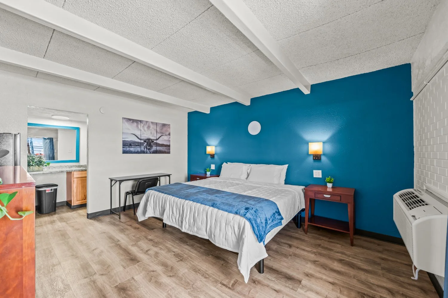 Hotel room with a bed against a blue accent wall, two nightstands with plants, wall-mounted lights, a small desk, and a kitchenette with a window, a framed cow mountain picture, and an air conditioning unit.
