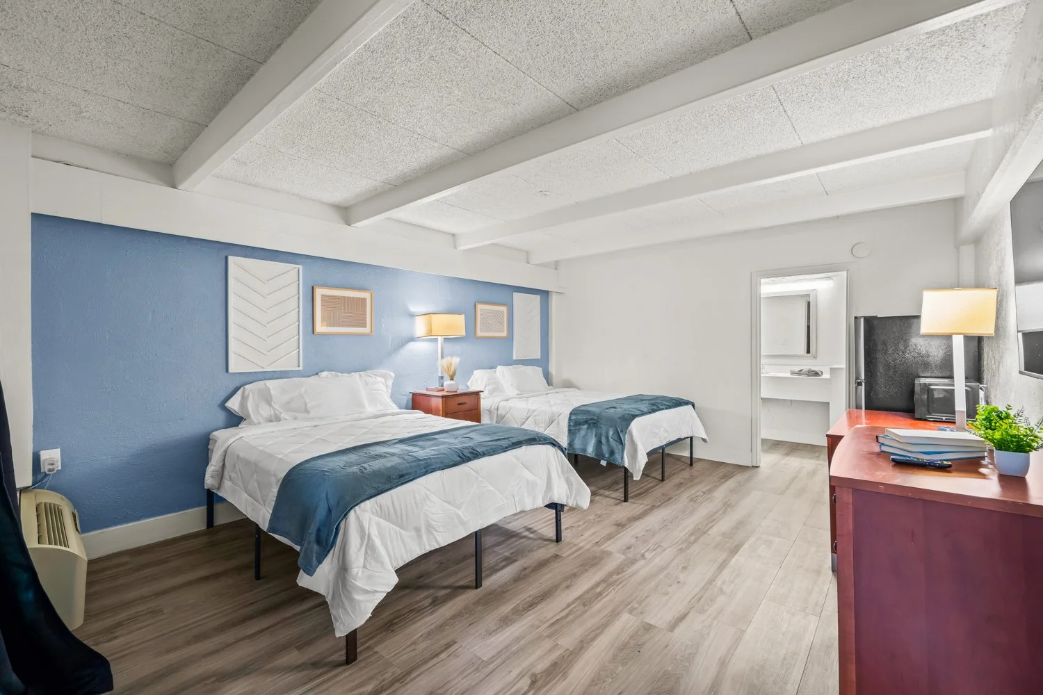 A hotel room with two beds, a nightstand with a lamp, a blue accent wall, and a desk with a TV, books, a lamp, and a plant.