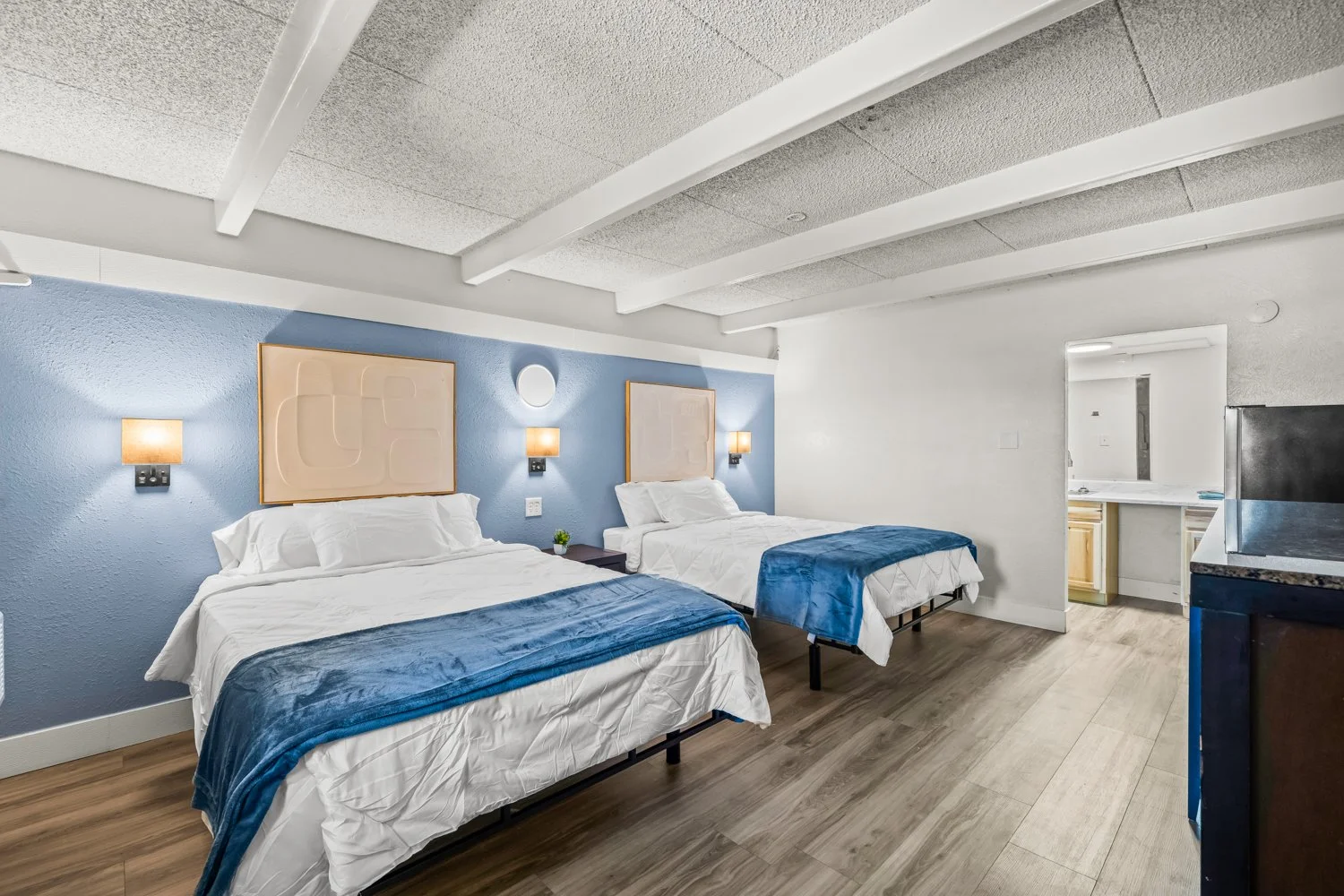 Hotel room with two beds with white linens and blue throws, blue accent wall, and wooden floors. There are wall-mounted lamps and framed artwork above each bed. A small envelope paper plant is between the beds. An open doorway reveals a small kitchen