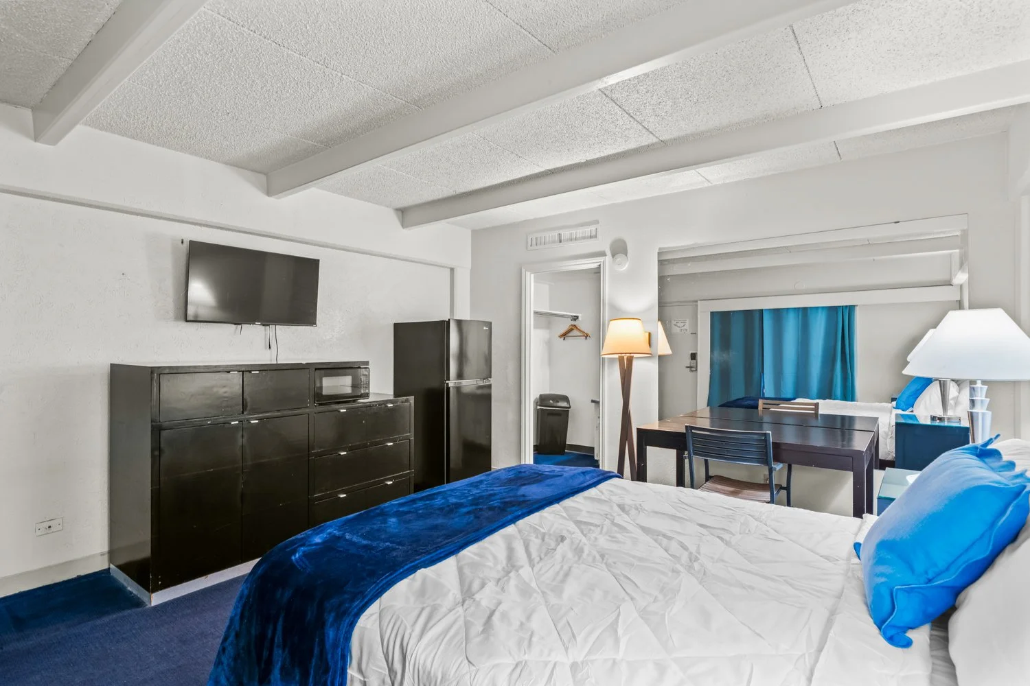 Hotel room with two beds, a dark wood dresser, a small refrigerator, a TV mounted on the wall, a nightstand, a lamp, a small dining table with chairs, a window with blue curtains, and a closet area with a hanger.