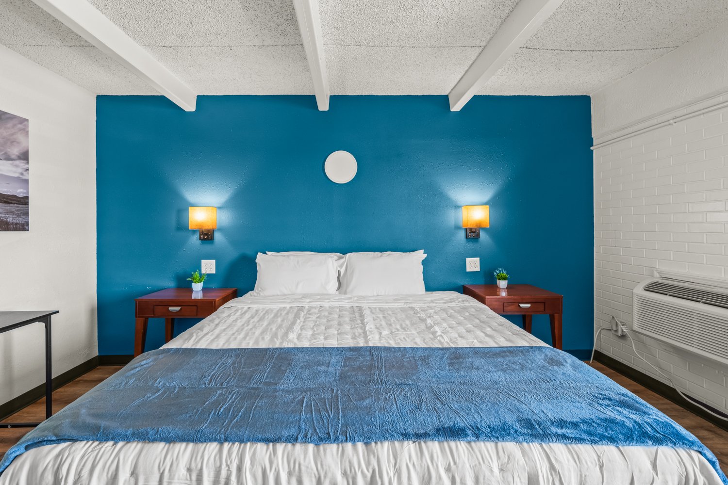 A neatly made bed with white pillows and a white comforter, has a blue blanket at the foot, in a room with a blue accent wall, two wooden nightstands with small potted plants, wall-mounted lamps, and a framed picture on the white wall.