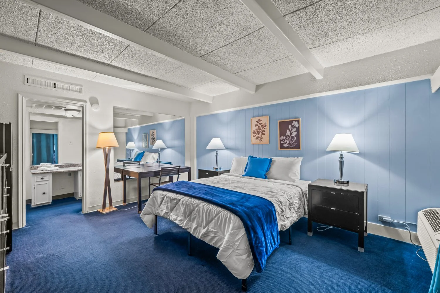 A hotel room with a bed covered in white bedding and blue accents, a black nightstand with a lamp, and blue wall paneling decorated with framed botanical prints.