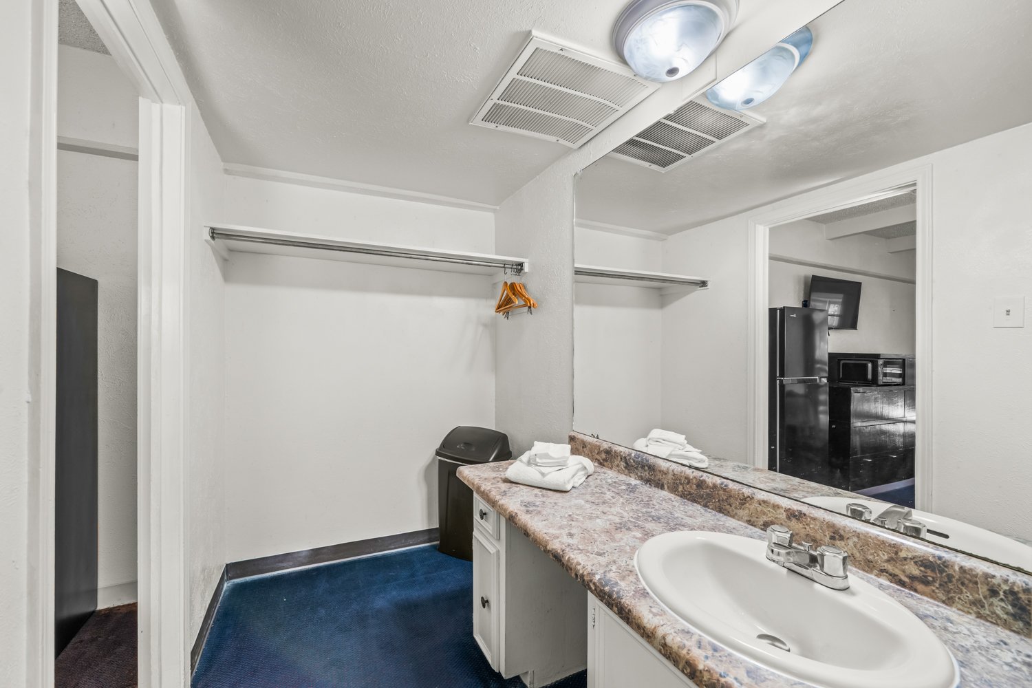 Empty hotel room bathroom with a sink, mirror, folded towels, and a small trash can, visible in the reflection of the mirror.