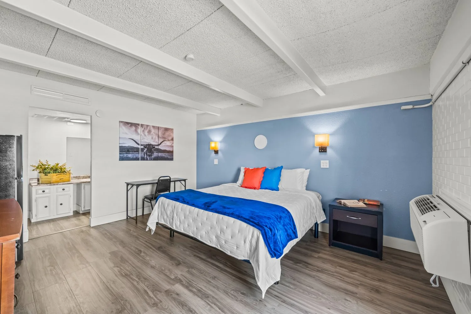 Hotel room with a bed, pillows, and a blue and white color scheme, including wall-mounted lamps and a small table with magazines.