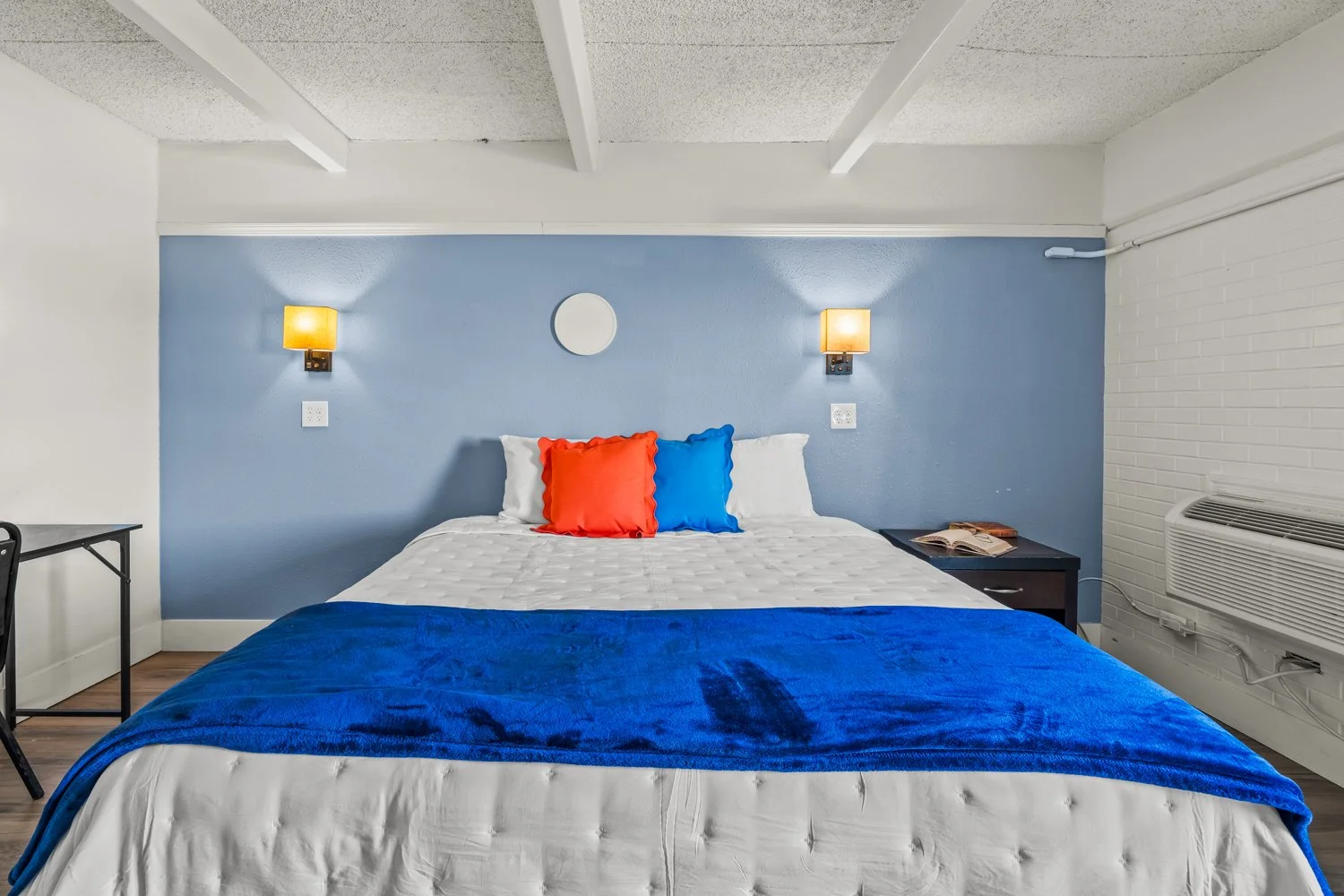 A neatly made bed with white bedding and two colorful pillows, one red and one blue, against a blue accent wall with two wall-mounted lamps.
