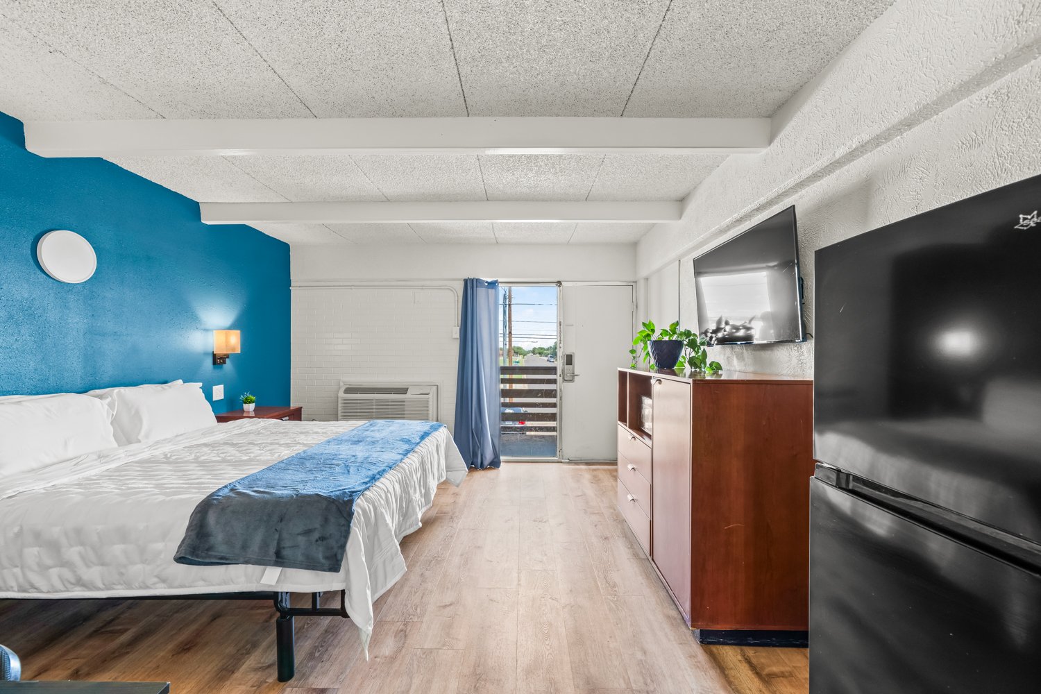 Interior of a motel room with a bed, nightstand, wall-mounted light, TV, dresser, refrigerator, plant, and a door leading to a balcony.