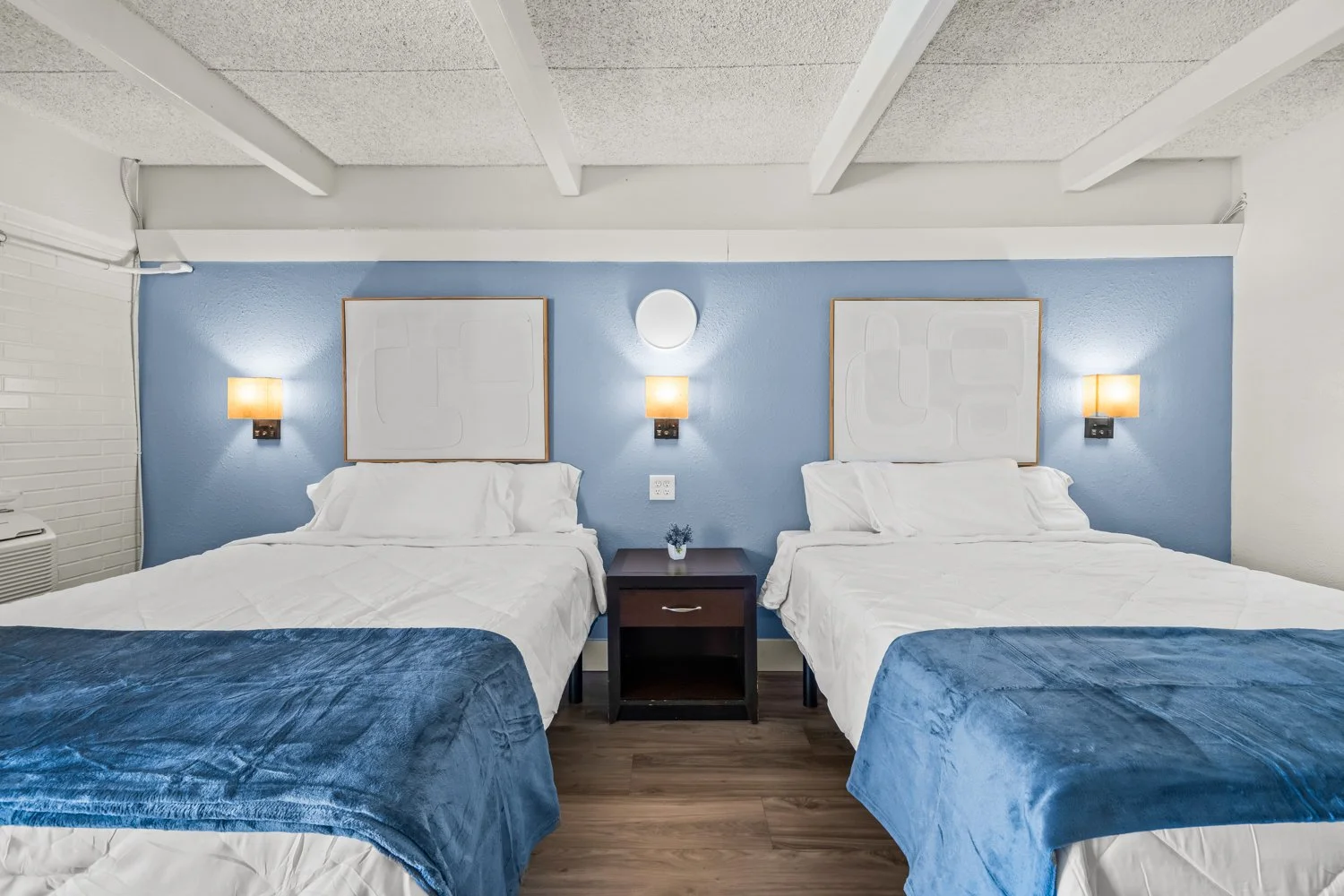A hotel room with two beds, each with blue blankets, white sheets, and pillows. There is a nightstand between the beds with a small purple plant. The wall behind the beds is light blue with two framed art pieces and mounted lights