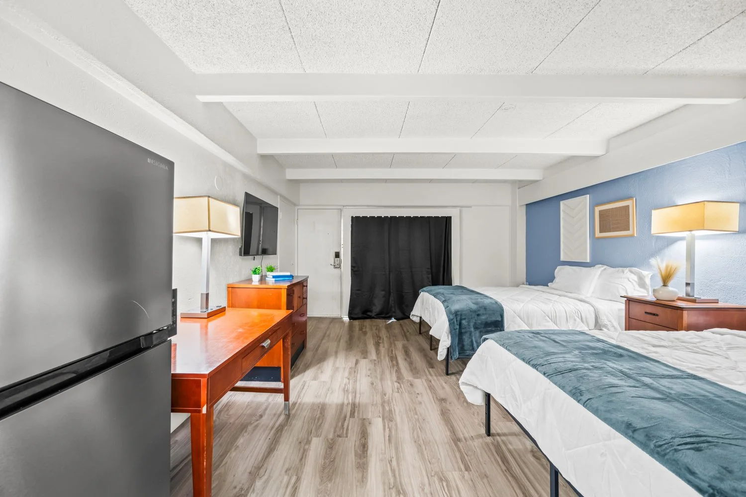 Interior of a hotel room with two beds, a flat-screen TV on a wooden dresser, a refrigerator, a small table, and wall-mounted lamps.