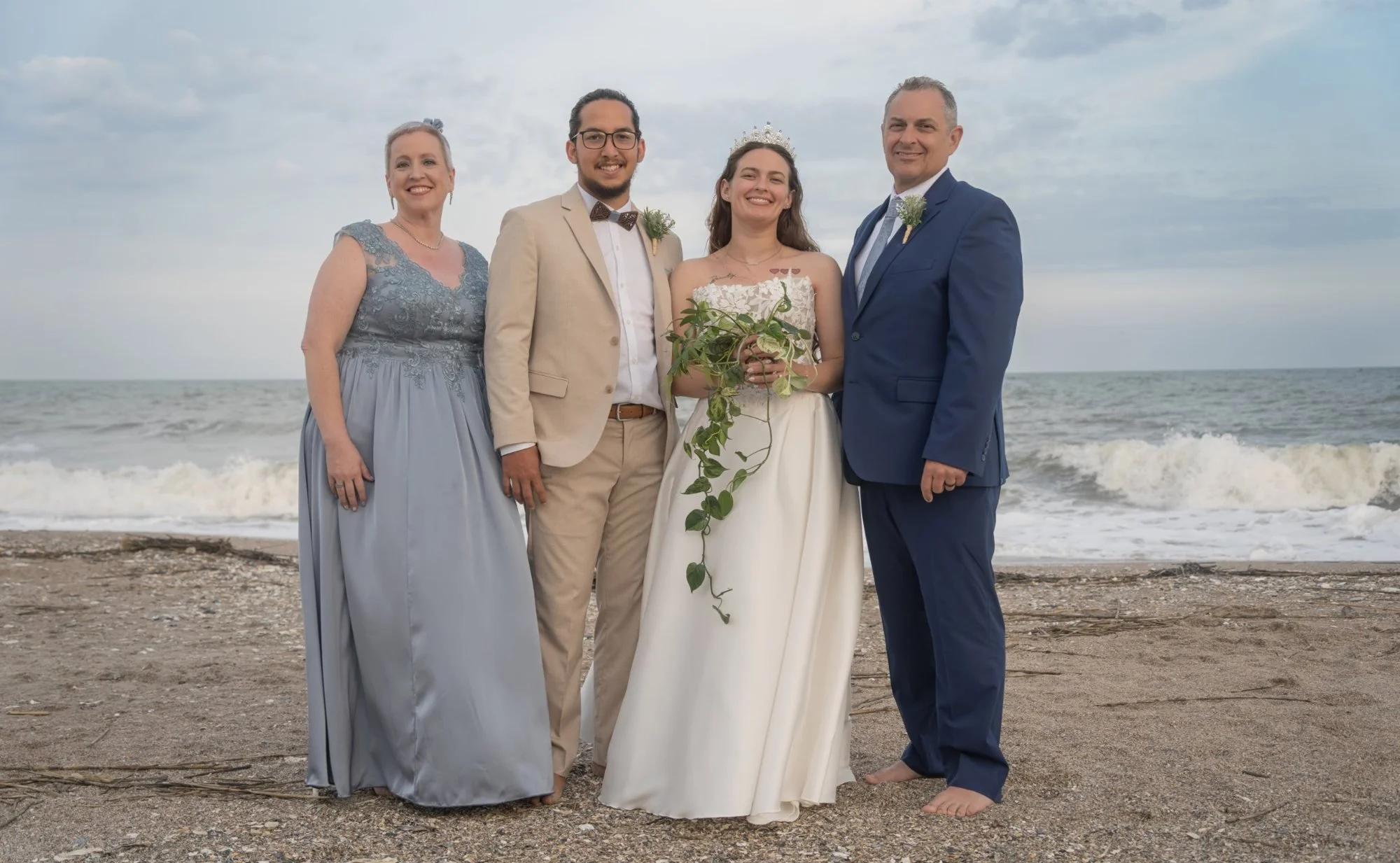 A group of five people standing on a beach during a wedding ceremony, with the ocean and cloudy sky in the background. The bride is wearing a white dress and holding a bouquet, the groom is in a beige suit with a bow tie, and the other three people a