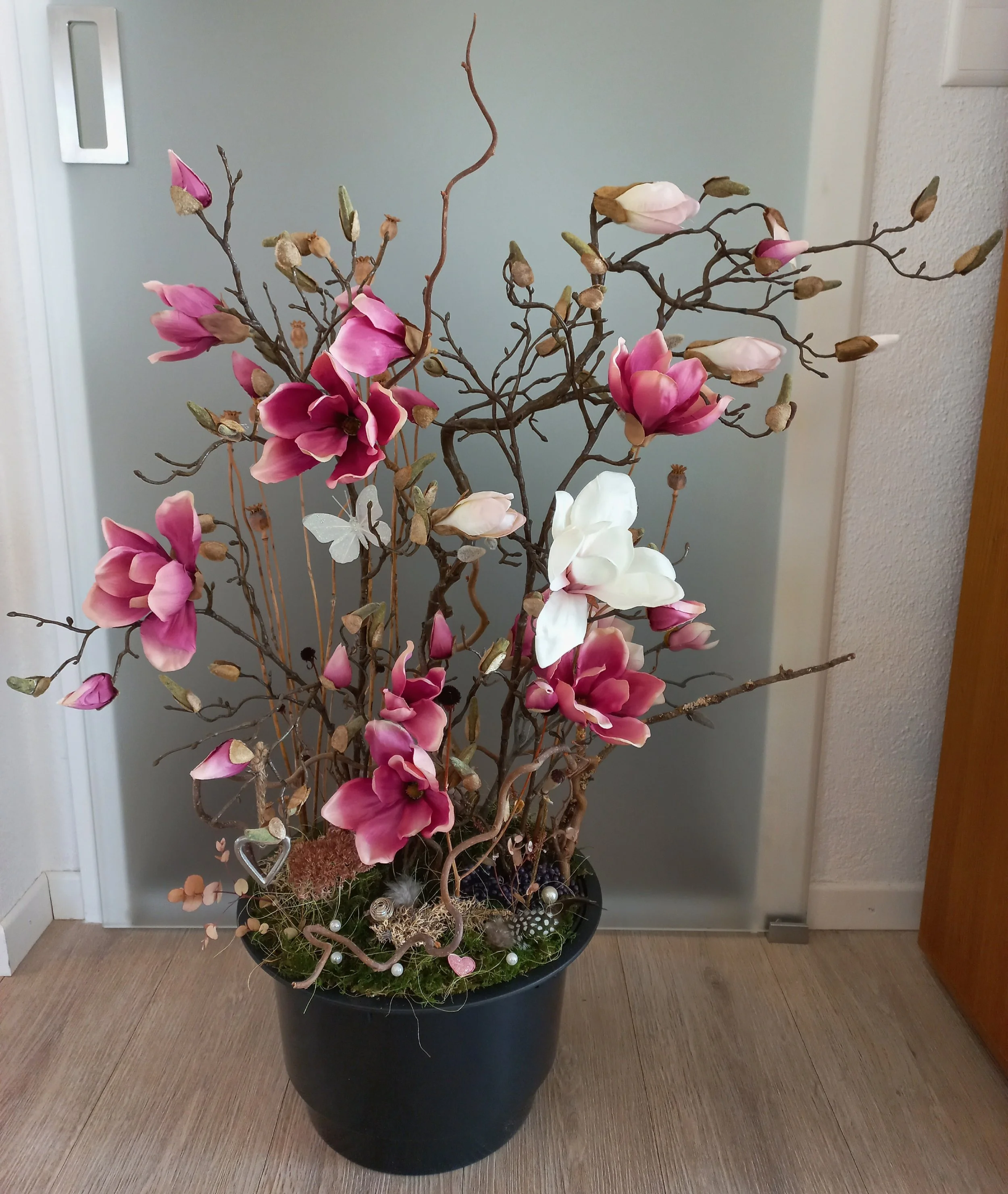 Kunstvoller Blütenstrauß in einer schwarzen Blumenvase mit rosa, weißen und lila Blüten, auf einem Holzboden vor einer grauen Wand.