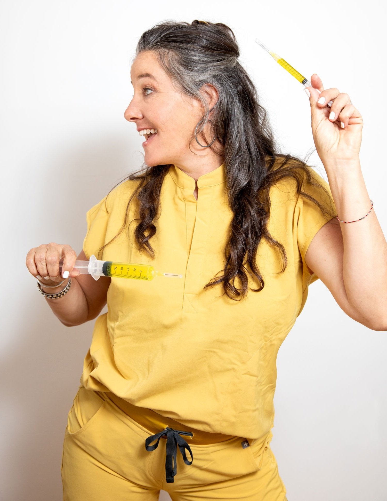 A woman holding two large medical syringes, one in each hand, smiling and looking to her left, against a plain white background.