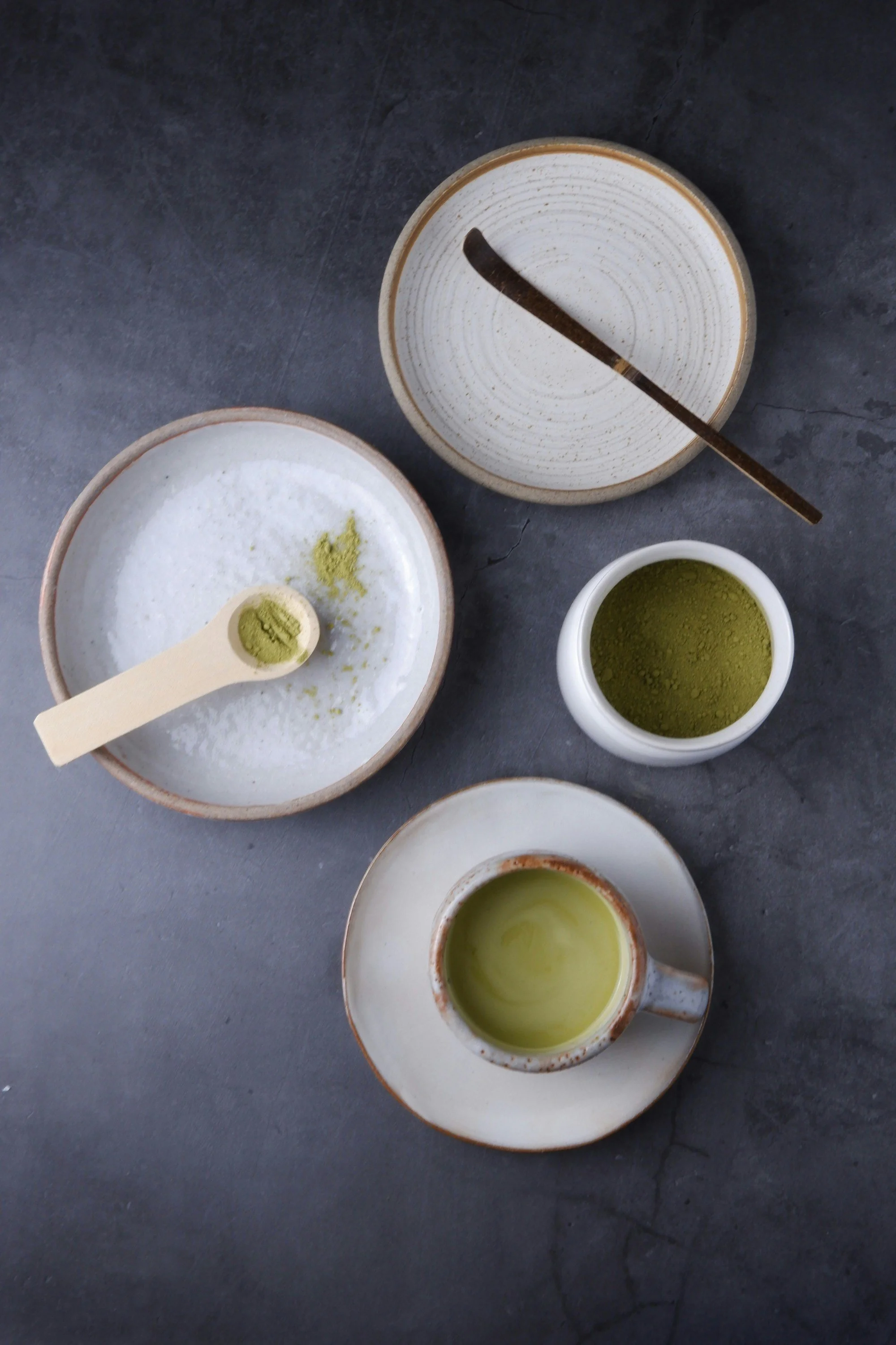Top view of matcha tea utensils including a cup of green tea, a bowl of powdered matcha with a scoop, an empty bowl with a stick, and a small jar of matcha powder.