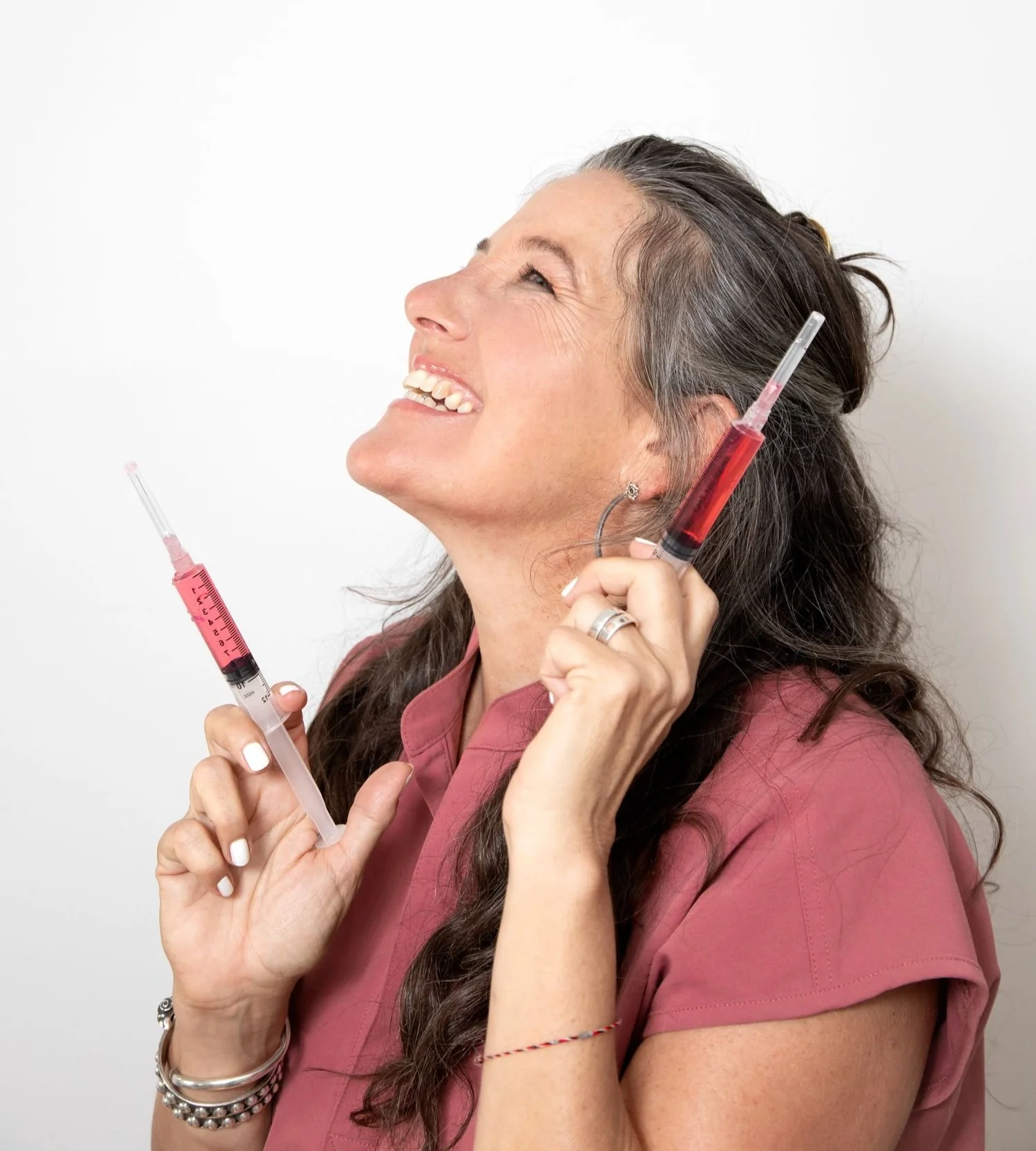 A woman holding multiple syringes filled with red and pink liquids, smiling and looking up