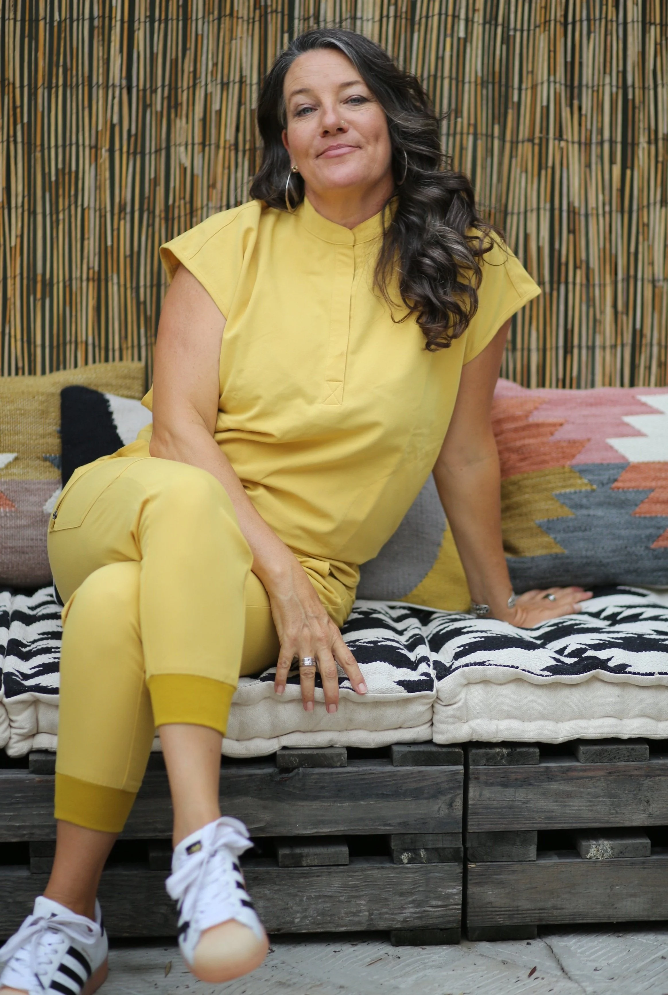 A woman with long dark hair wearing a yellow outfit sitting on a cushion with a zebra print, against a bamboo fence background, looking at the camera with a relaxed expression.
