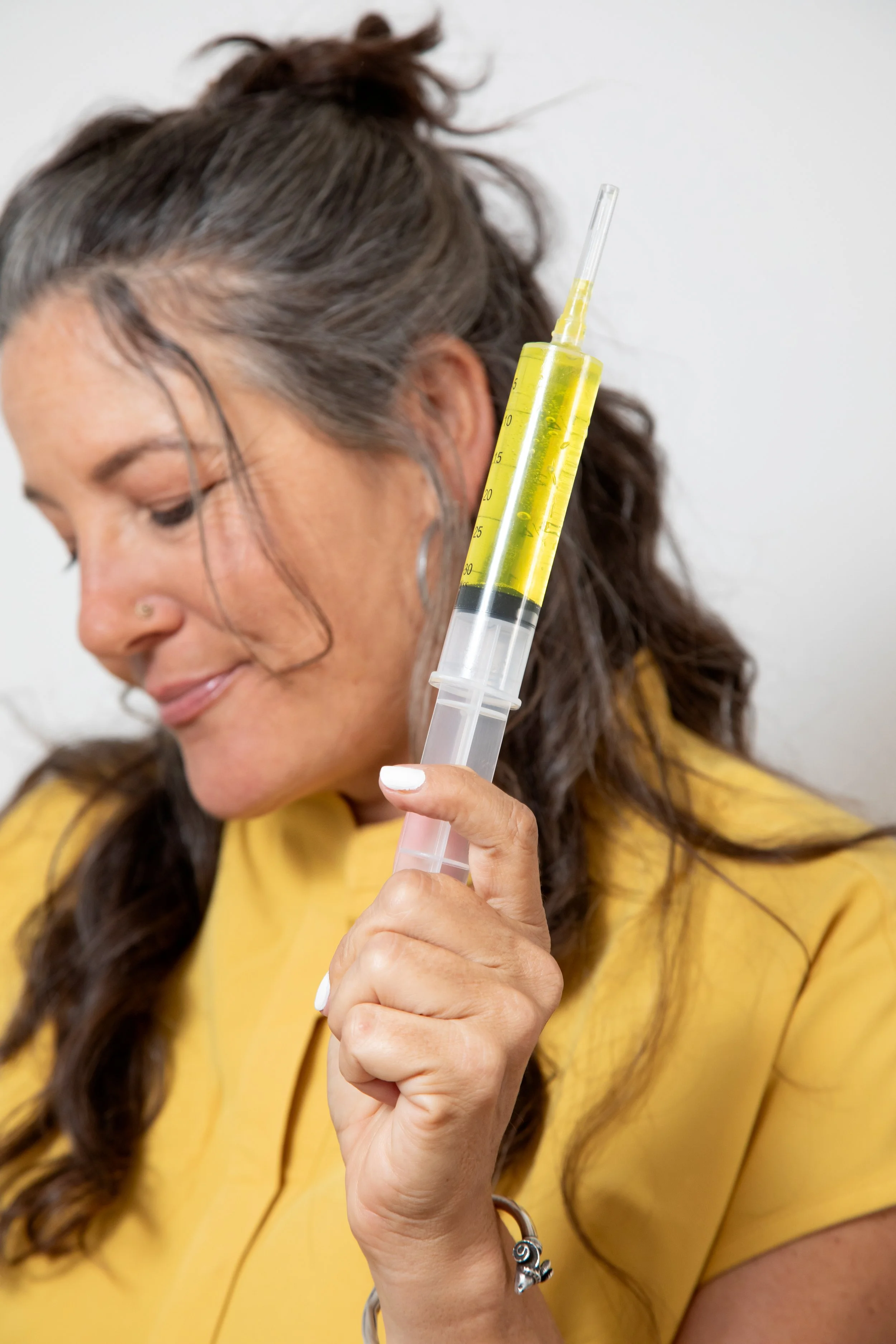 A woman with dark hair holding a large syringe filled with yellow liquid, close to her face, wearing a yellow shirt.