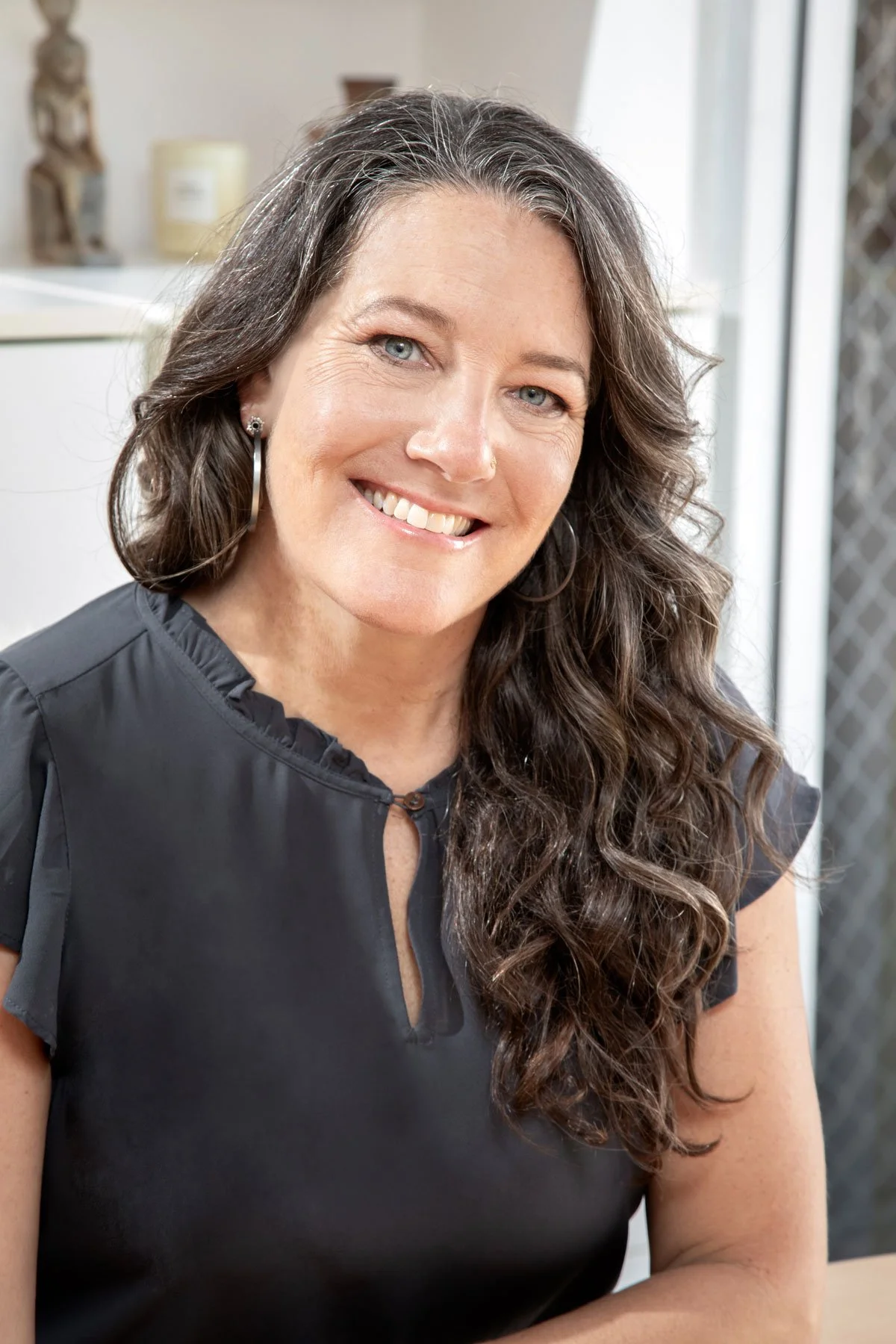 A woman with long, wavy brown hair and blue eyes, smiling and wearing a black top with ruffled edges near the neckline, sitting inside a room near a window.