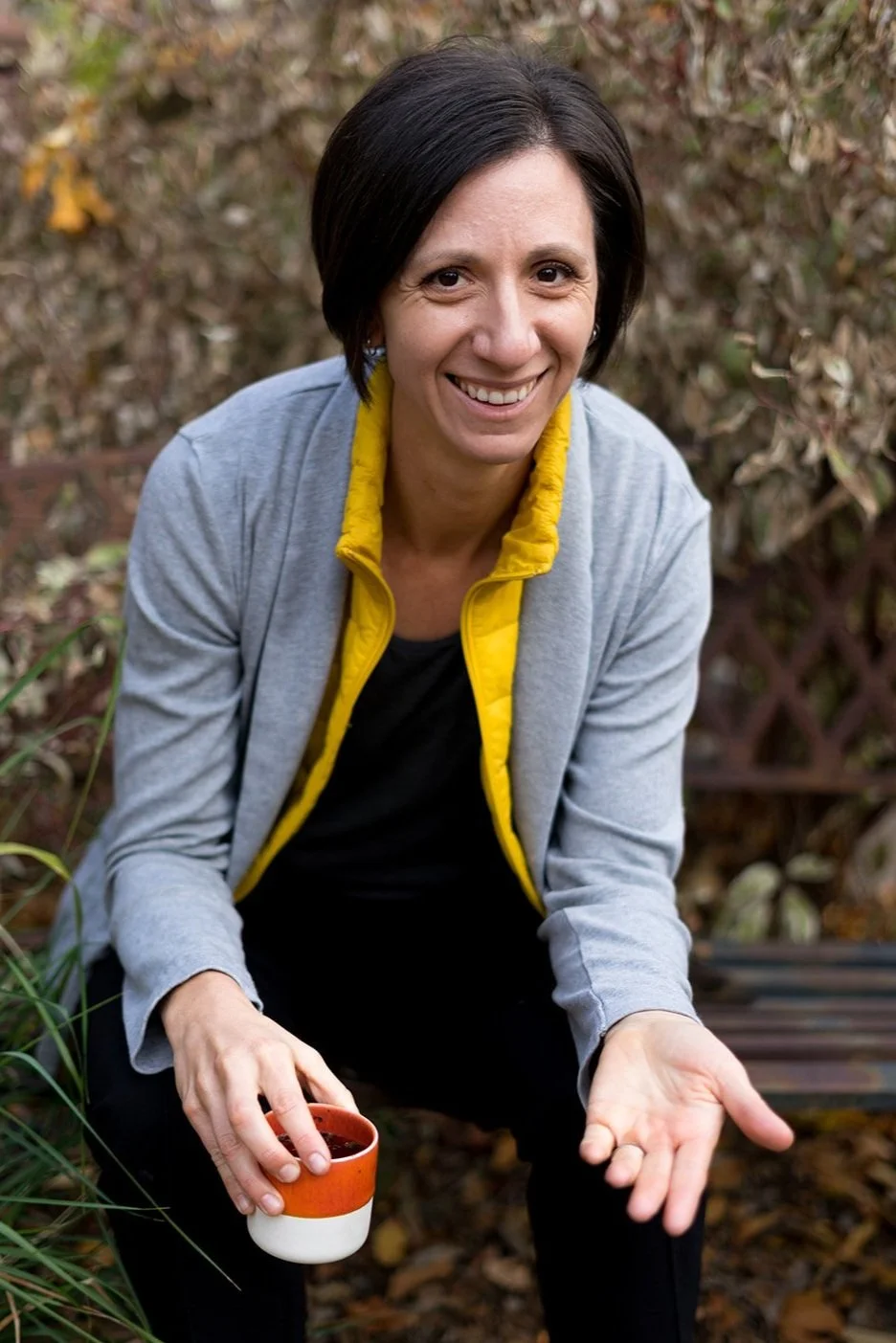 A naturopathic or functional medicine female doctor with short dark hair smiling outdoors, wearing a gray jacket with a yellow lining, holding a ceramic mug, with a background of autumn-colored foliage.