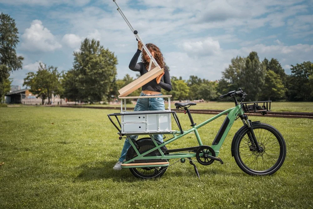 Eine Frau mit lockigem Haar steht an einem grünen E-Bike mit Ladefläche im Park, hebt ein Holzstück an.