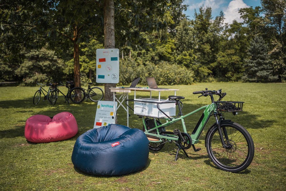 Ein Outdoor-Arbeitsbereich mit Fahrrädern, Sitzsäcken, Laptops, einem Whiteboard und einem Tisch im Park.