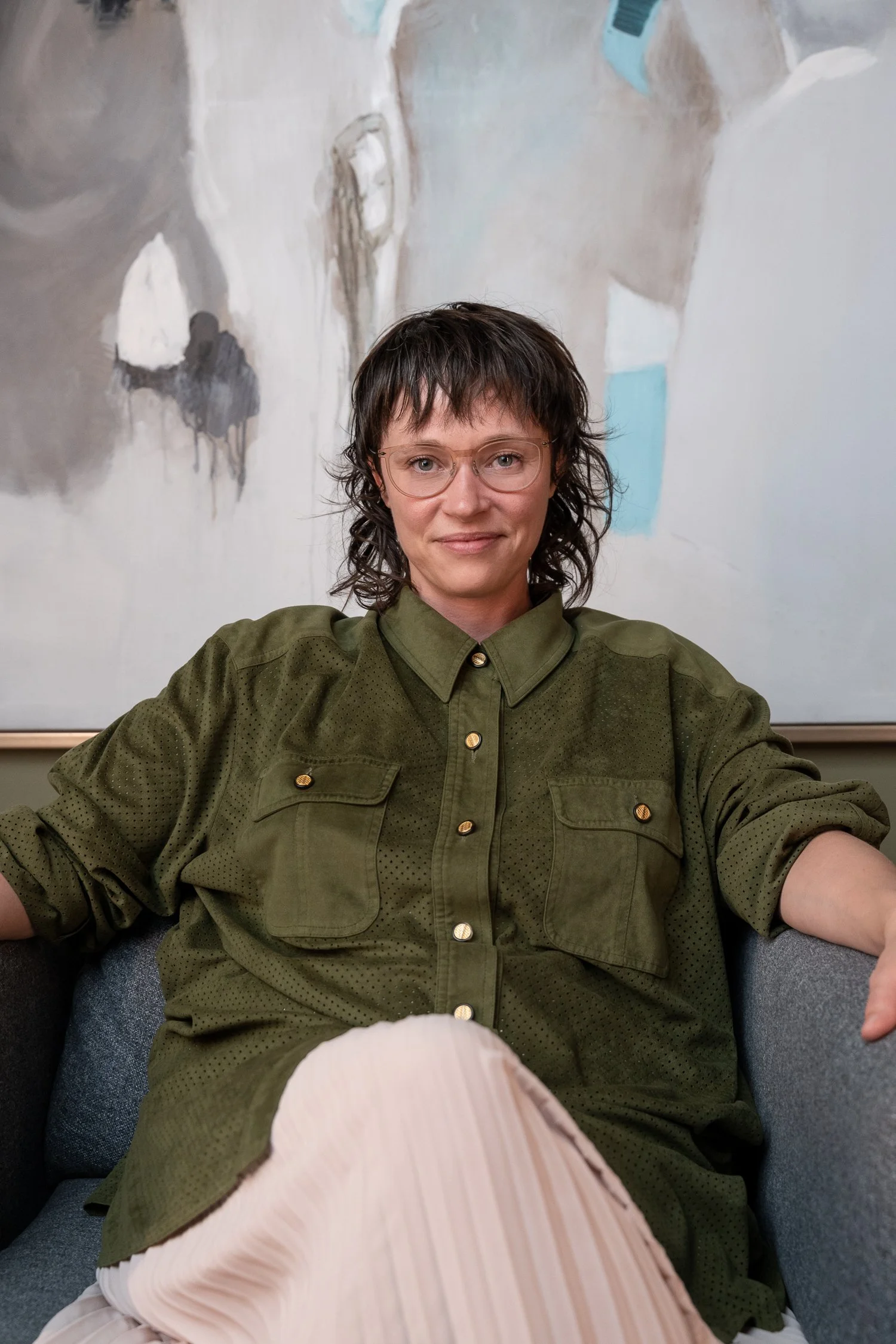 A person with glasses and shoulder-length dark hair, wearing an olive green button-up shirt sitting on a gray couch, with abstract artwork on the wall behind them.