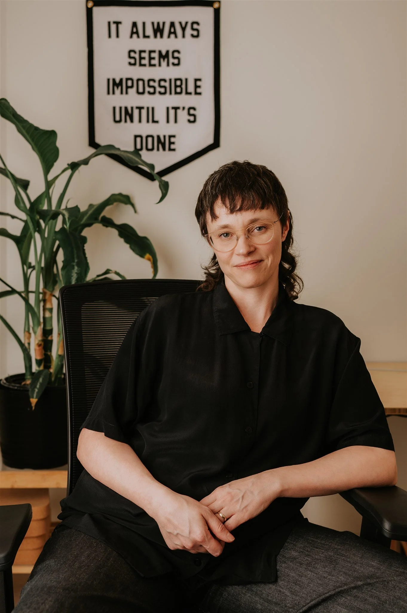 A person with glasses and shoulder-length dark hair, wearing an olive green button-up shirt sitting on a gray couch, with abstract artwork on the wall behind them.