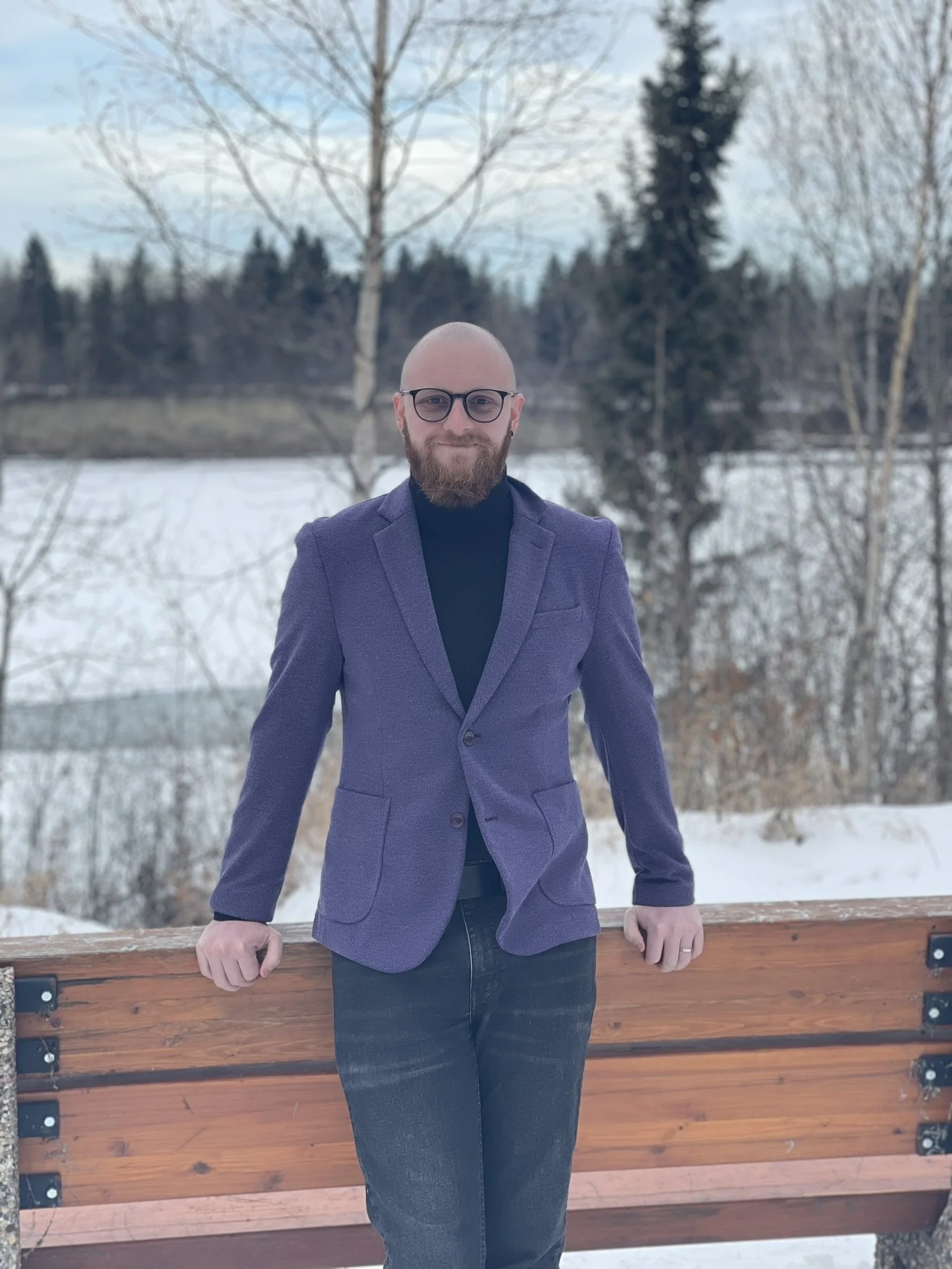 A man with a beard and glasses standing outdoors on a wooden bench with a snowy landscape and trees in the background.