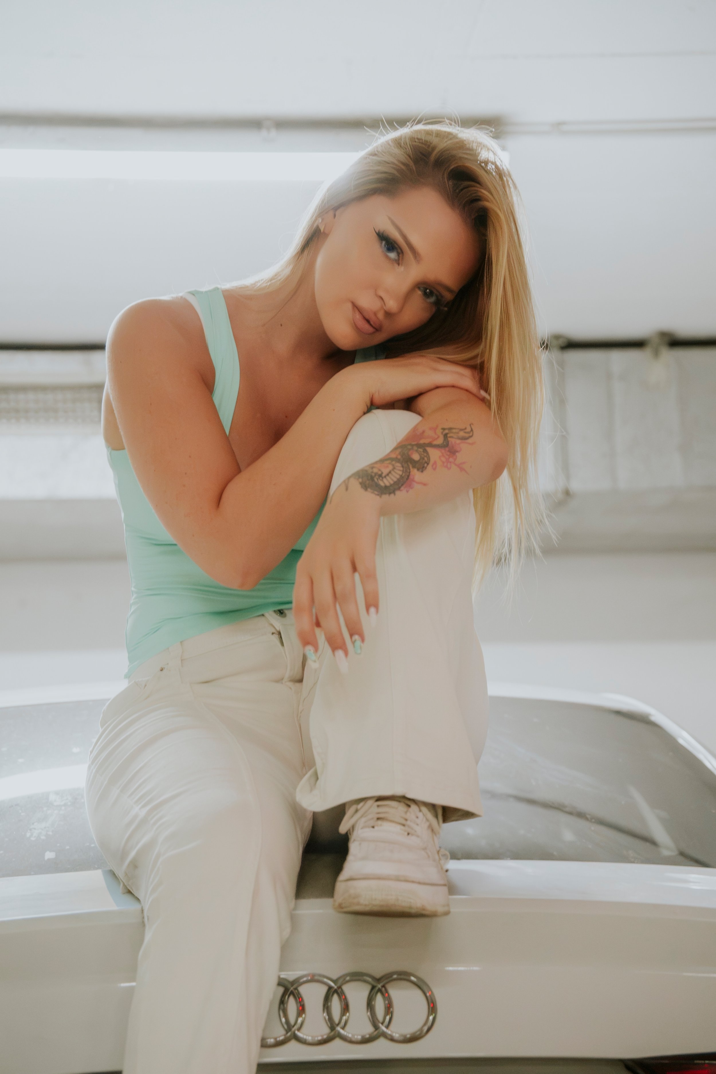 Junge Frau mit blonden Haaren, tätowiertem Arm, in einem hellblauen Top, sitzt auf einer weißen Oberfläche in einem Raum mit weißem Hintergrund, neben einem Auto mit sichtbarem Audi-Logo.