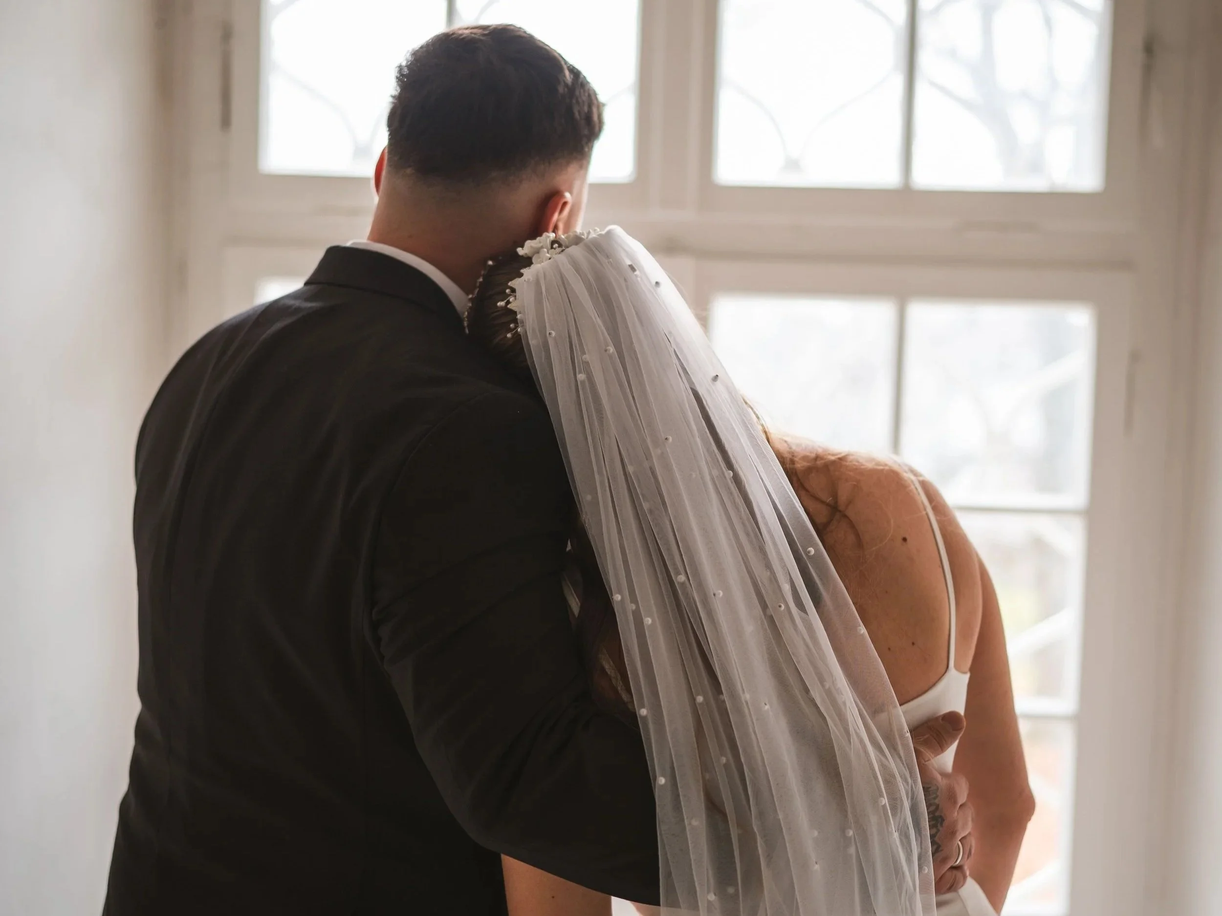 Ein Mann und eine Frau, die sich bei einer Hochzeit umarmen, vor einem großen Fenster mit natürlichem Licht.