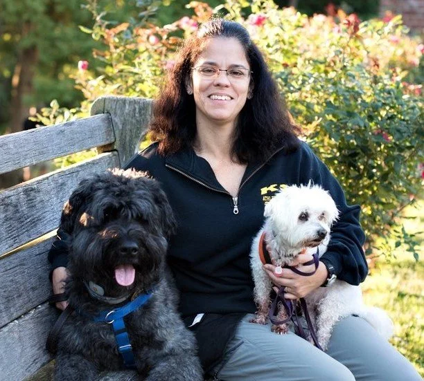 Woman sitting on outdoor park bench with two dogs, surrounded by greenery and flowers.
