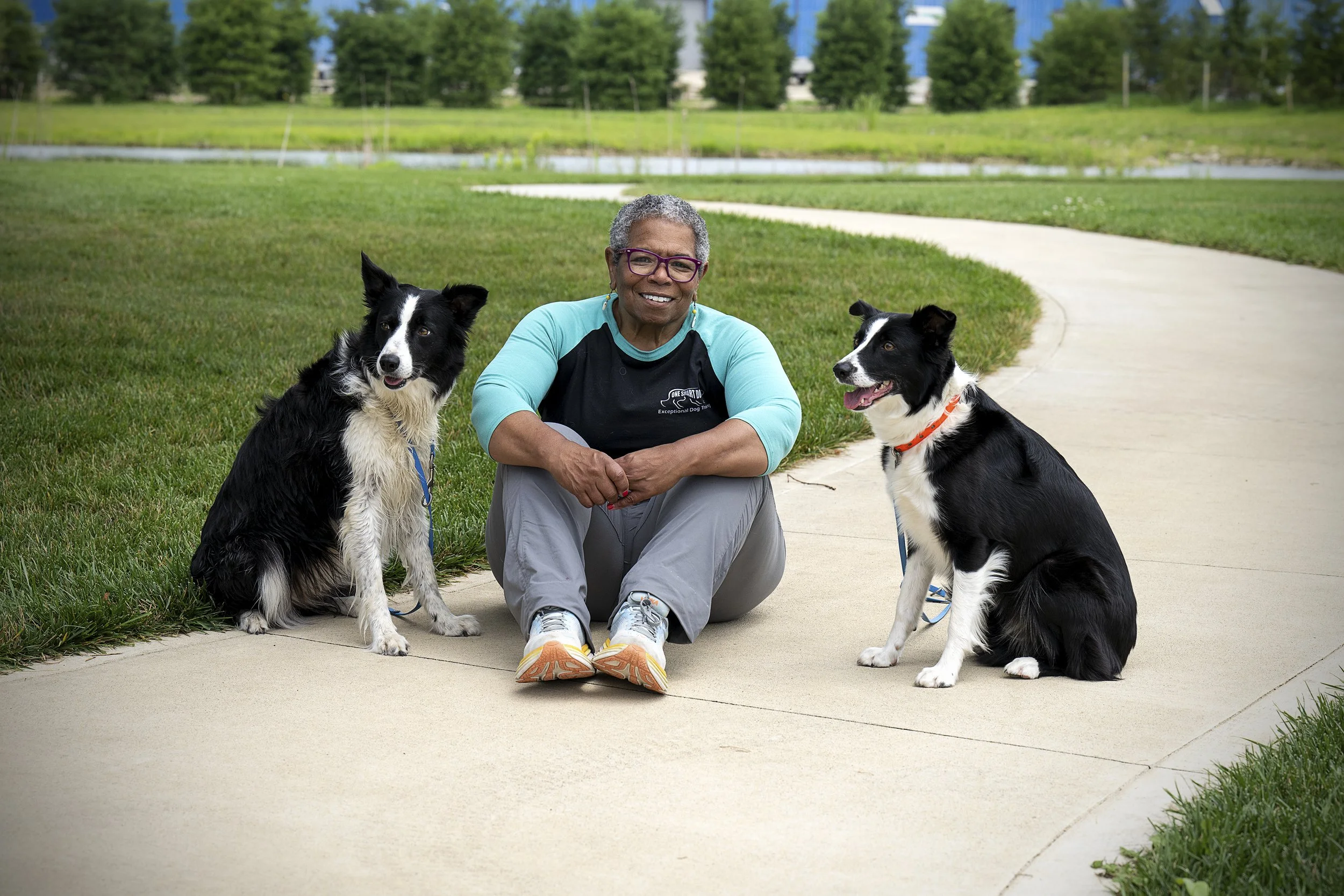 A smiling woman with gray hair, glasses, and a turquoise and black long-sleeve shirt sitting on a sidewalk with two black and white Border Collie dogs. The woman is posing in a park with green grass, trees, and a pond in the background.