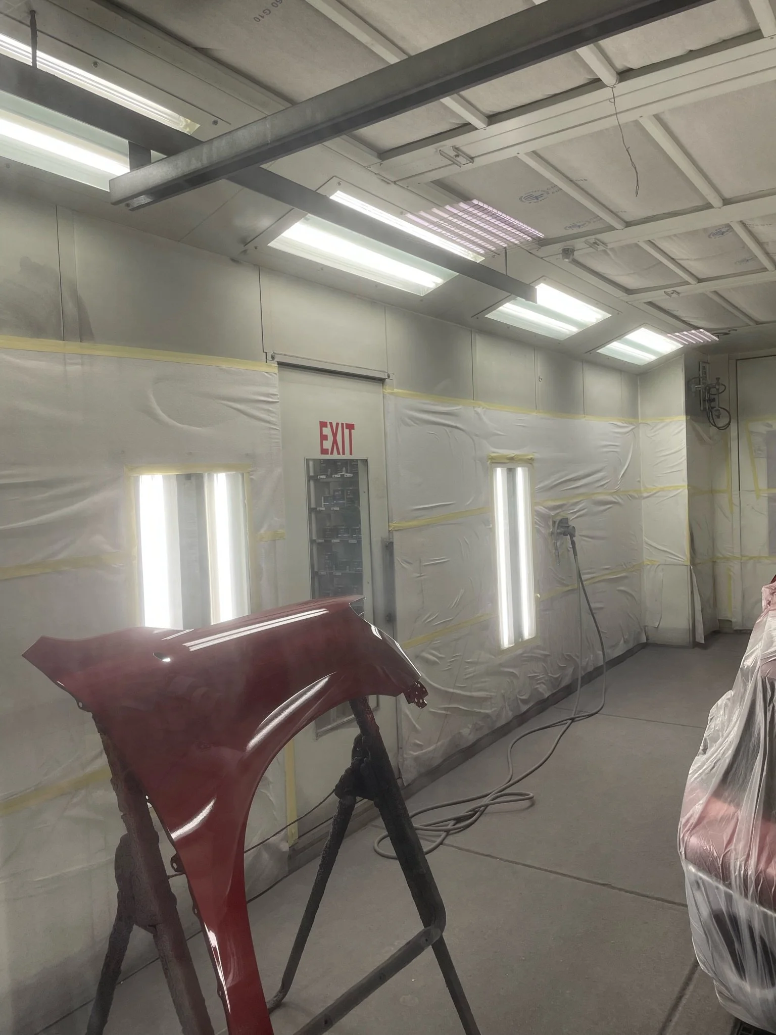 An automotive paint booth with fluorescent lighting, a red car fender on a stand, and a red exit sign on the wall.
