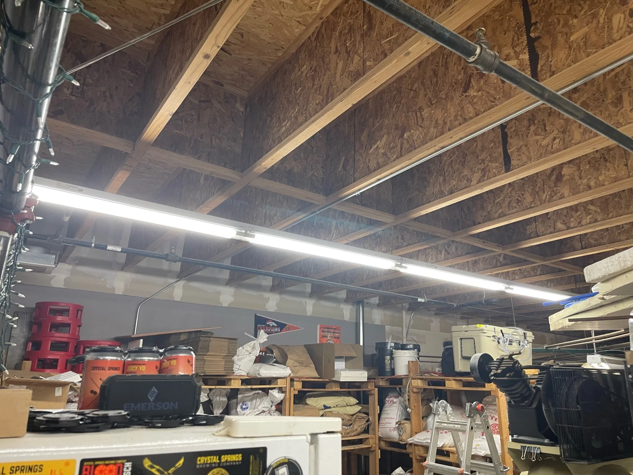 Garage with wooden beams, fluorescent lights, storage shelves filled with boxes, containers, and supplies, and an aluminum step ladder.